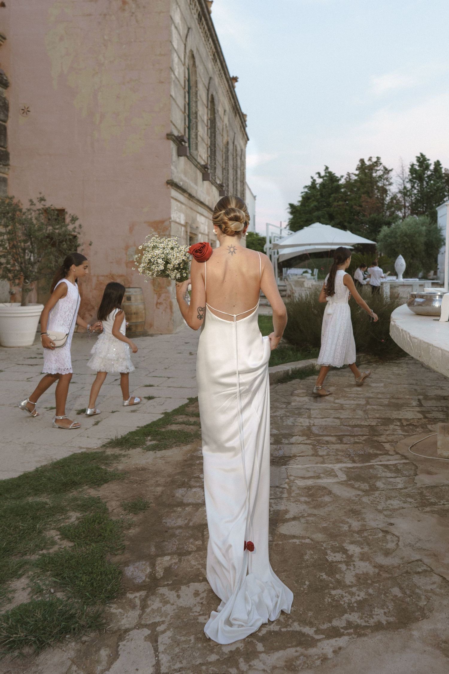A wedding scene outdoors with a person in an elegant white backless dress holding a bouquet of white flowers.