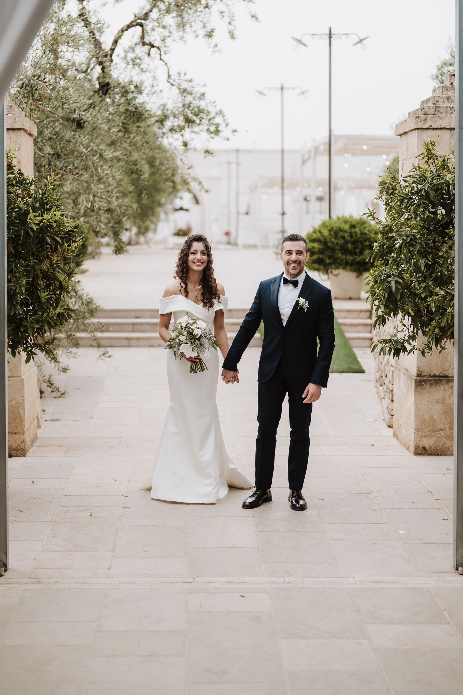 A couple in formal wedding attire stands together on a walkway with water views in the background.
