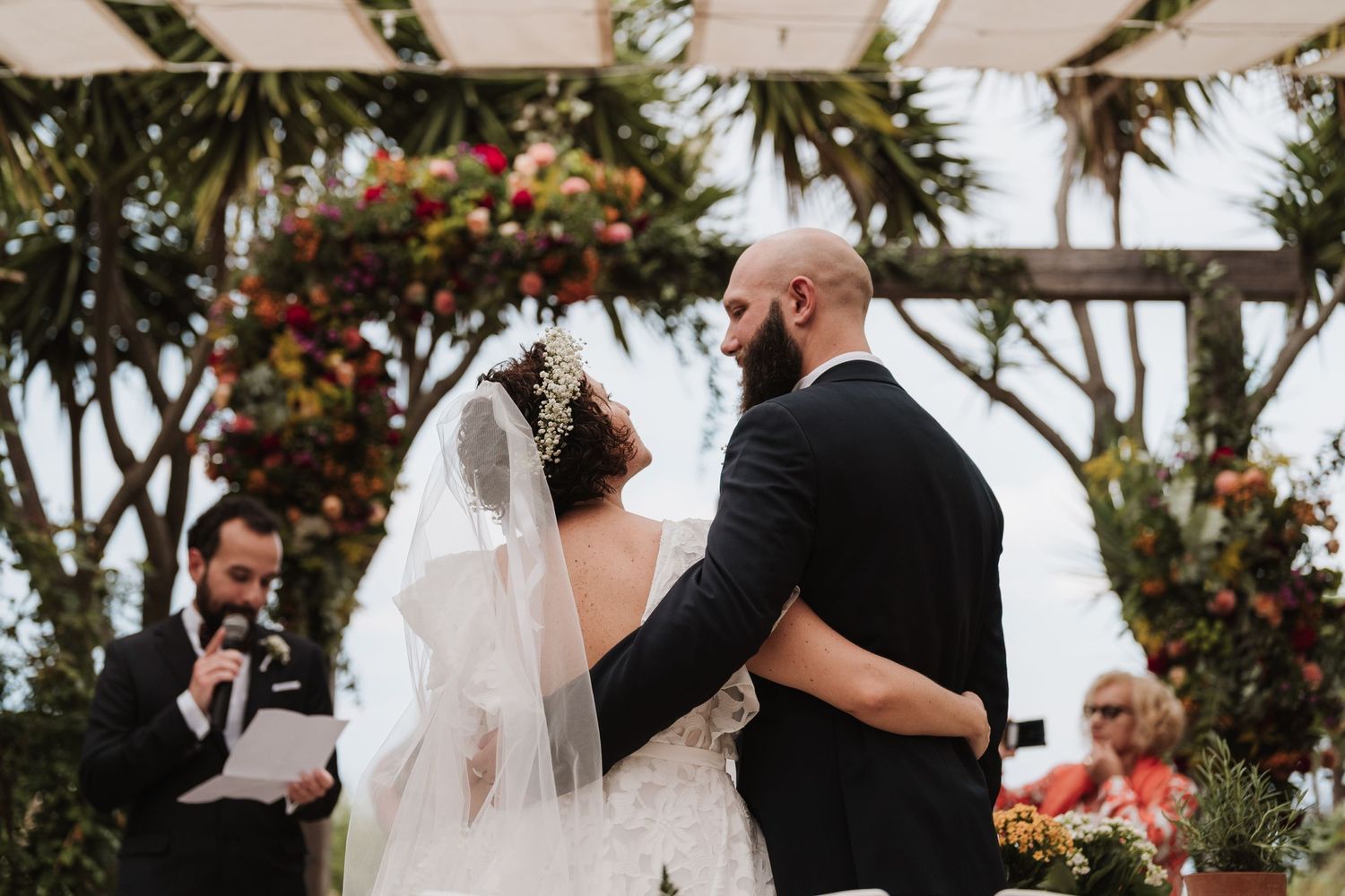 Outdoor wedding ceremony under a floral-decorated pergola with palm trees in the background.