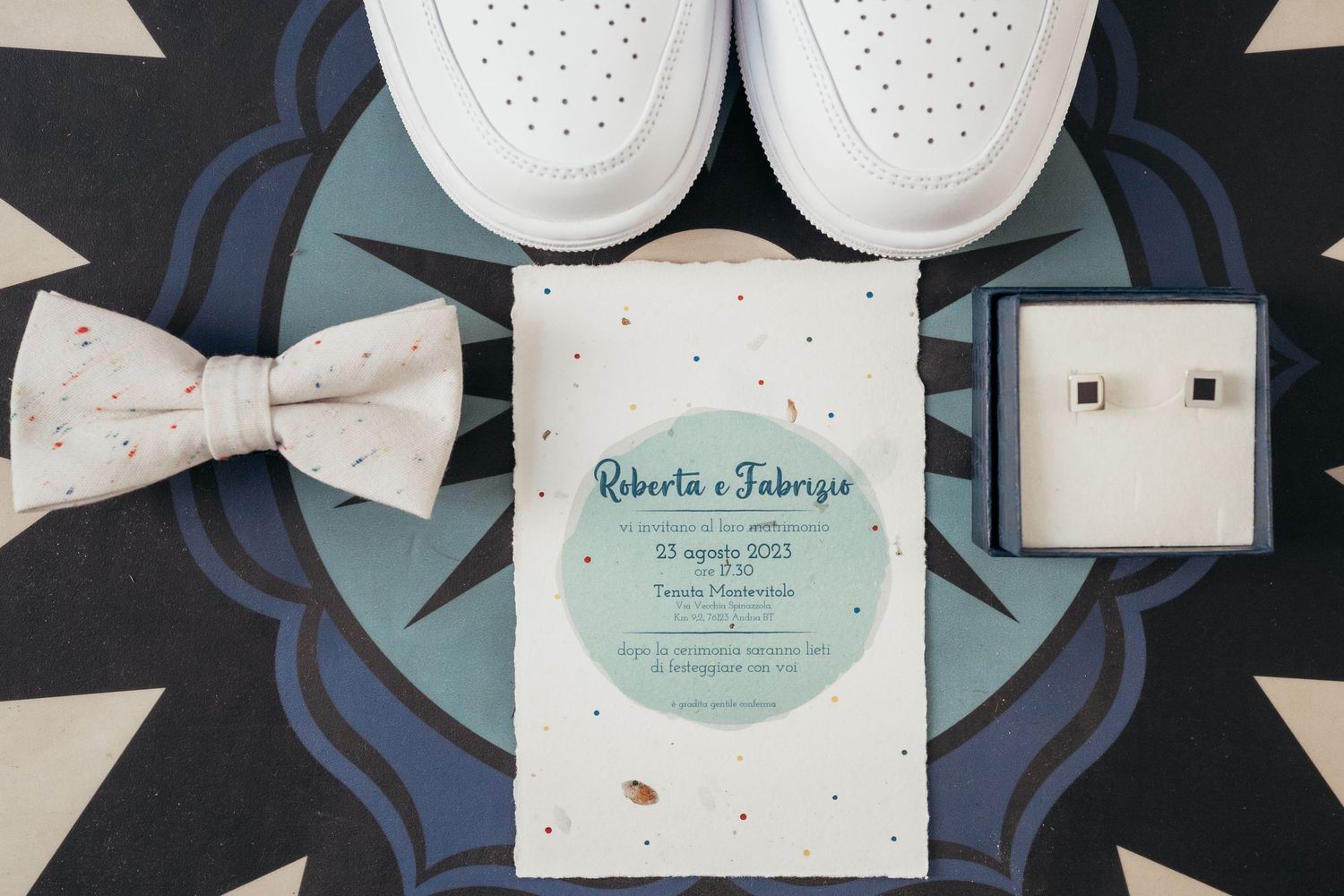 Wedding accessories including white shoes, bow tie, invitation and jewelry box arranged on patterned surface.