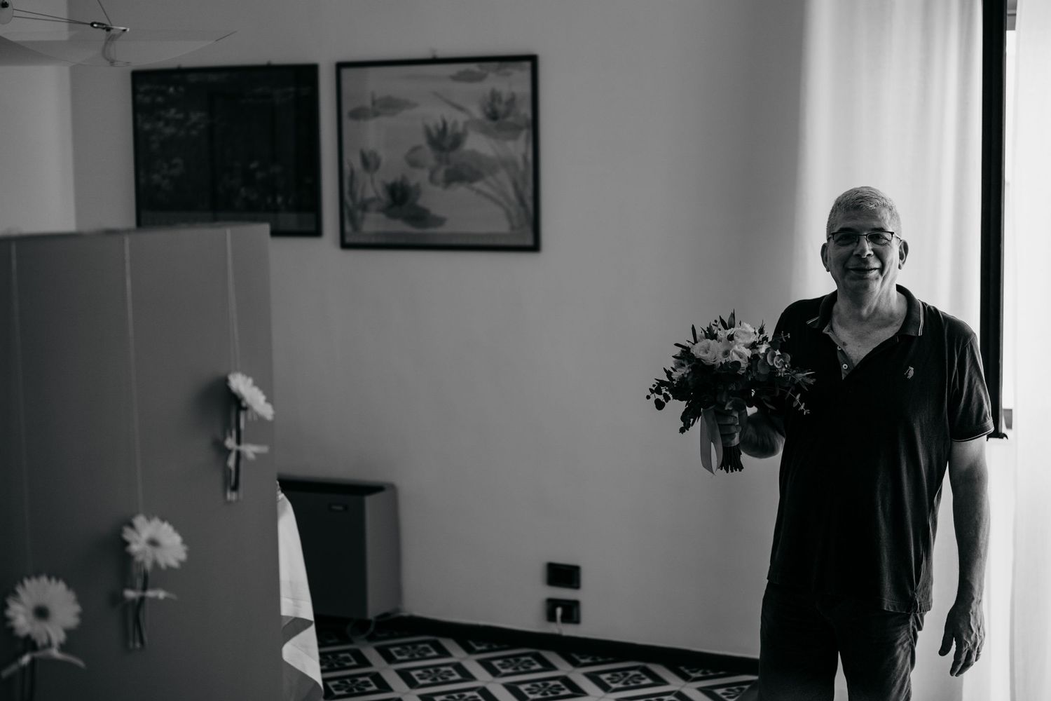 Black and white photo of someone holding a floral bouquet in a bedroom setting.