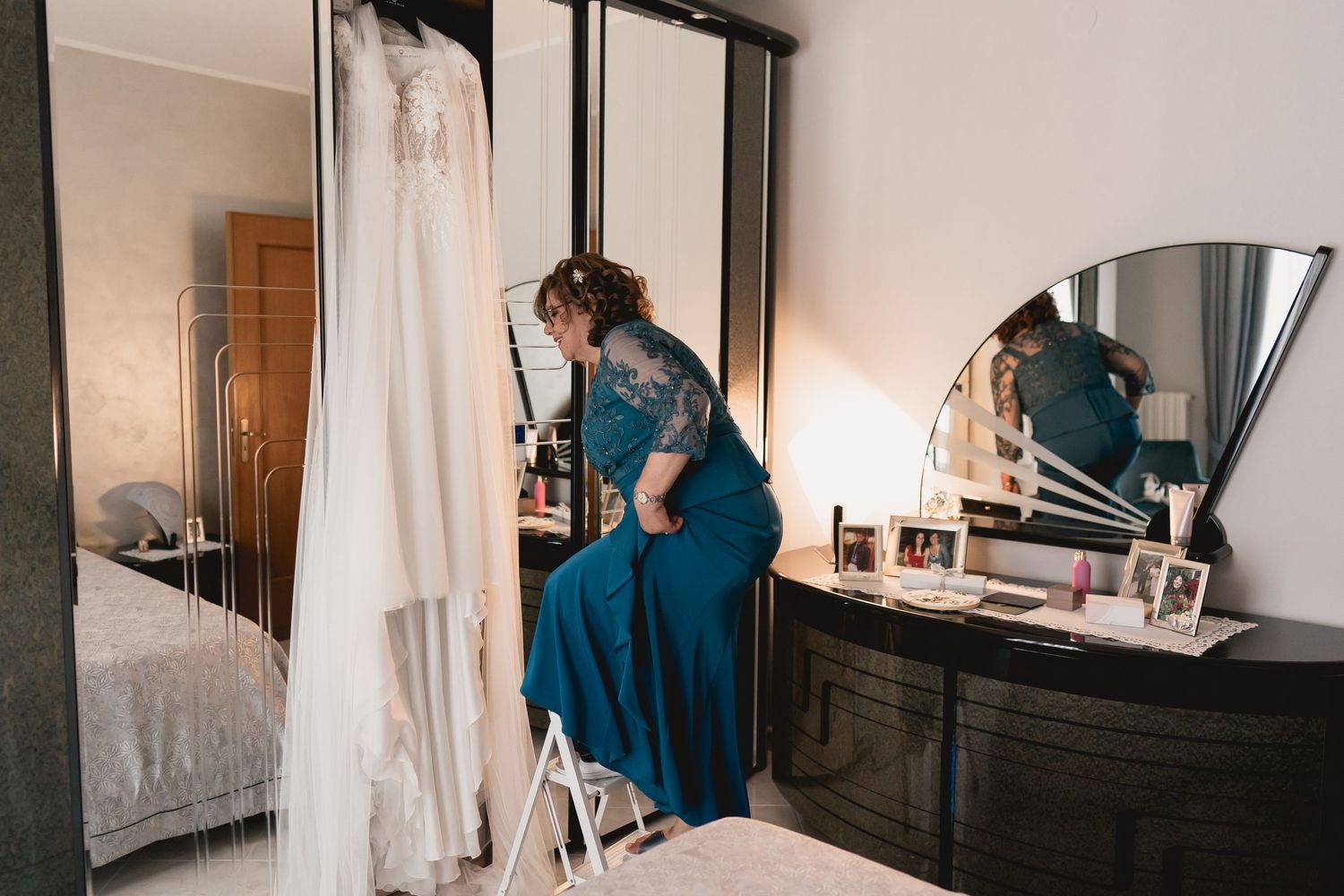 A woman in a blue dress stands on a ladder near a white wedding gown hanging in a bedroom.