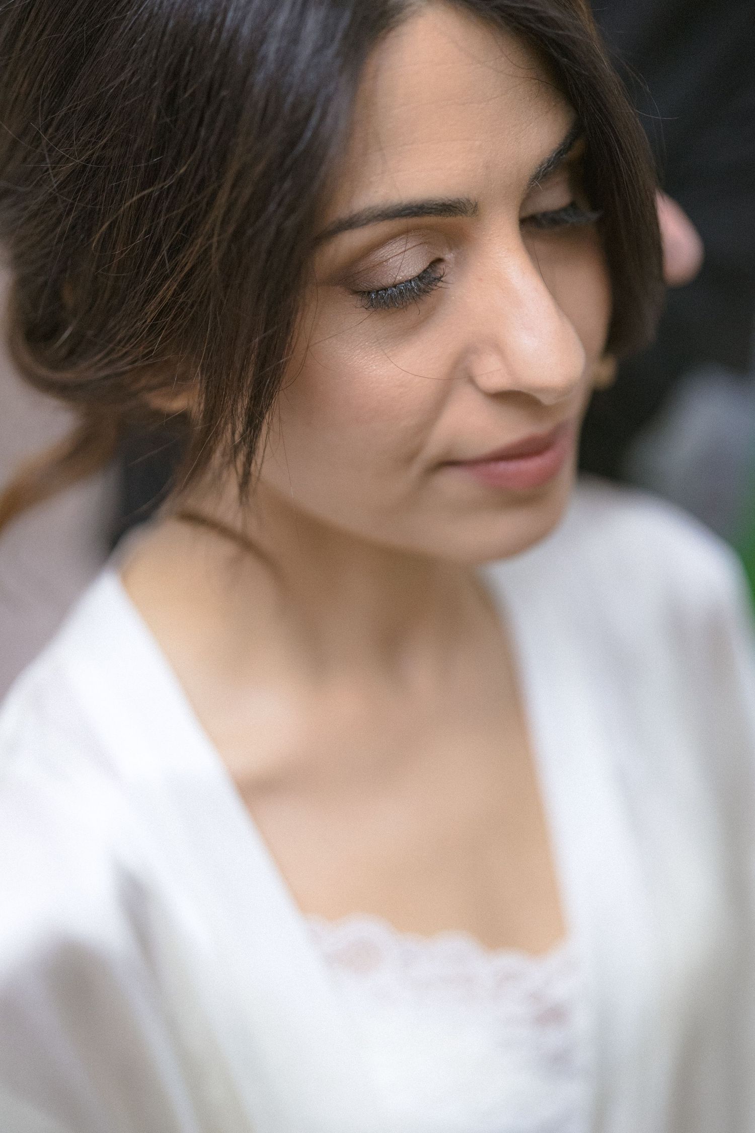 A bride in a white satin robe with lace detail looks down serenely during wedding preparations.