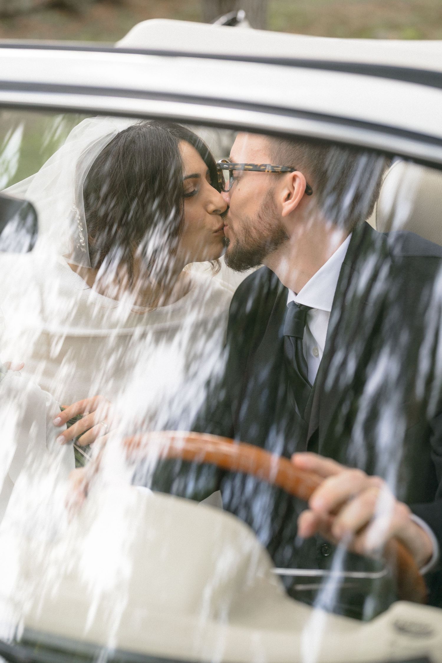 Bride and groom sharing a romantic kiss inside a vintage car on their wedding day.