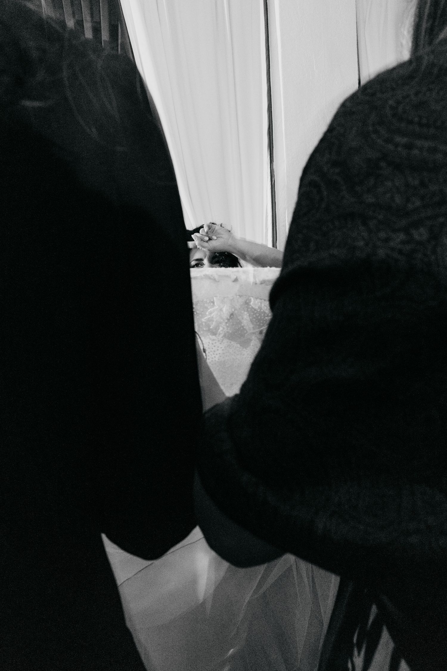 Two guests frame bride in white wedding dress visible through gap in black and white photo.