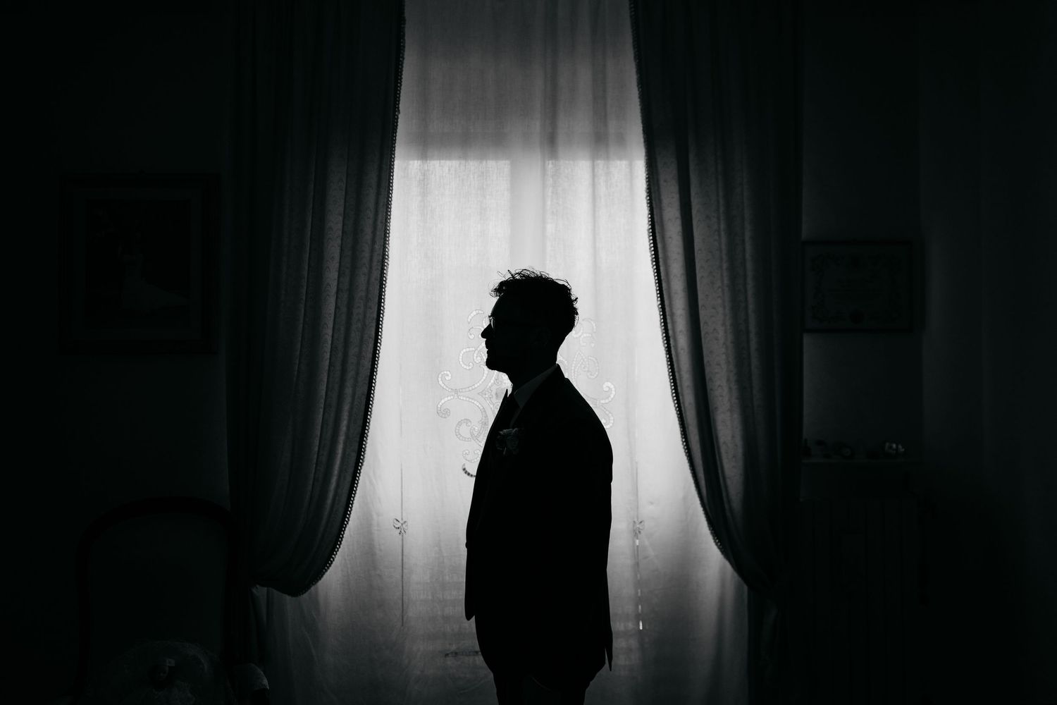 Silhouette of a man standing alone by a curtained window in a dark room.
