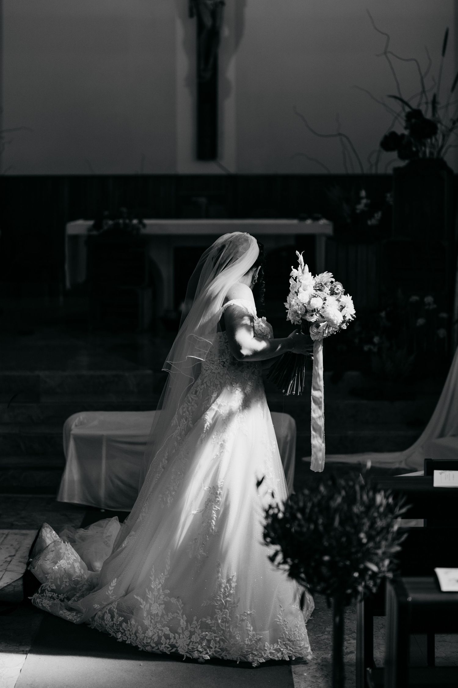 Black and white photo of bride in lace gown kneeling with bouquet inside a church.
