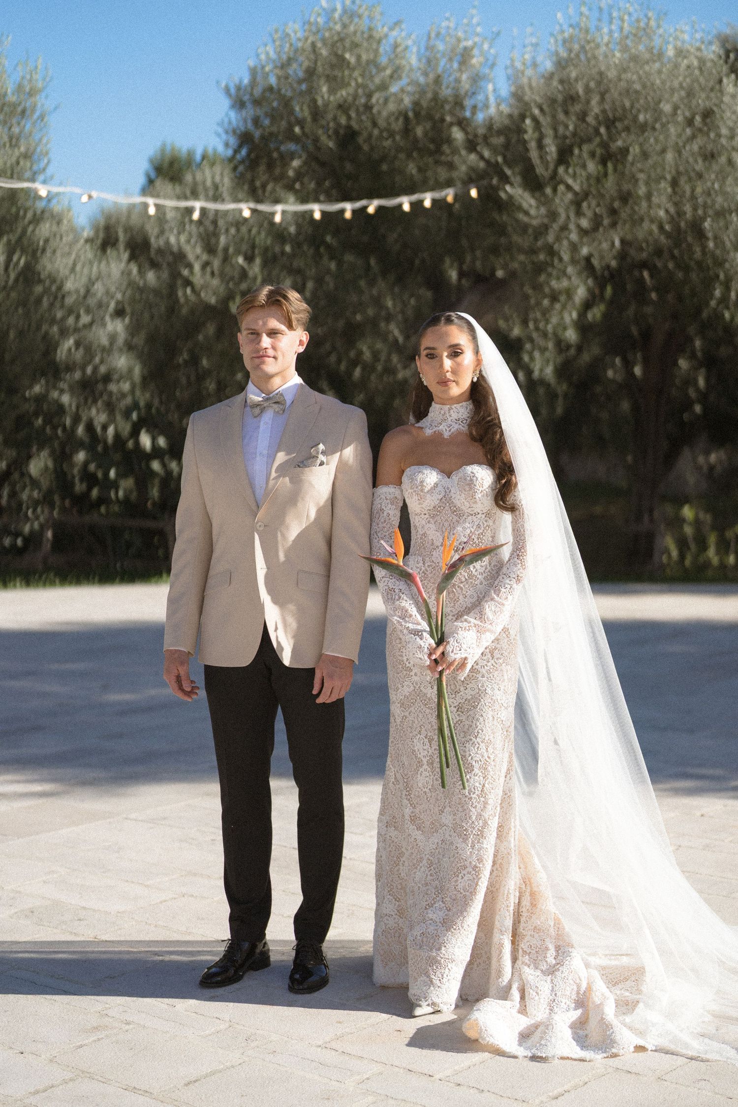 Bride and groom in a beige blazer and bow tie stand together on a sunny terrace beneath olive trees and string lights.