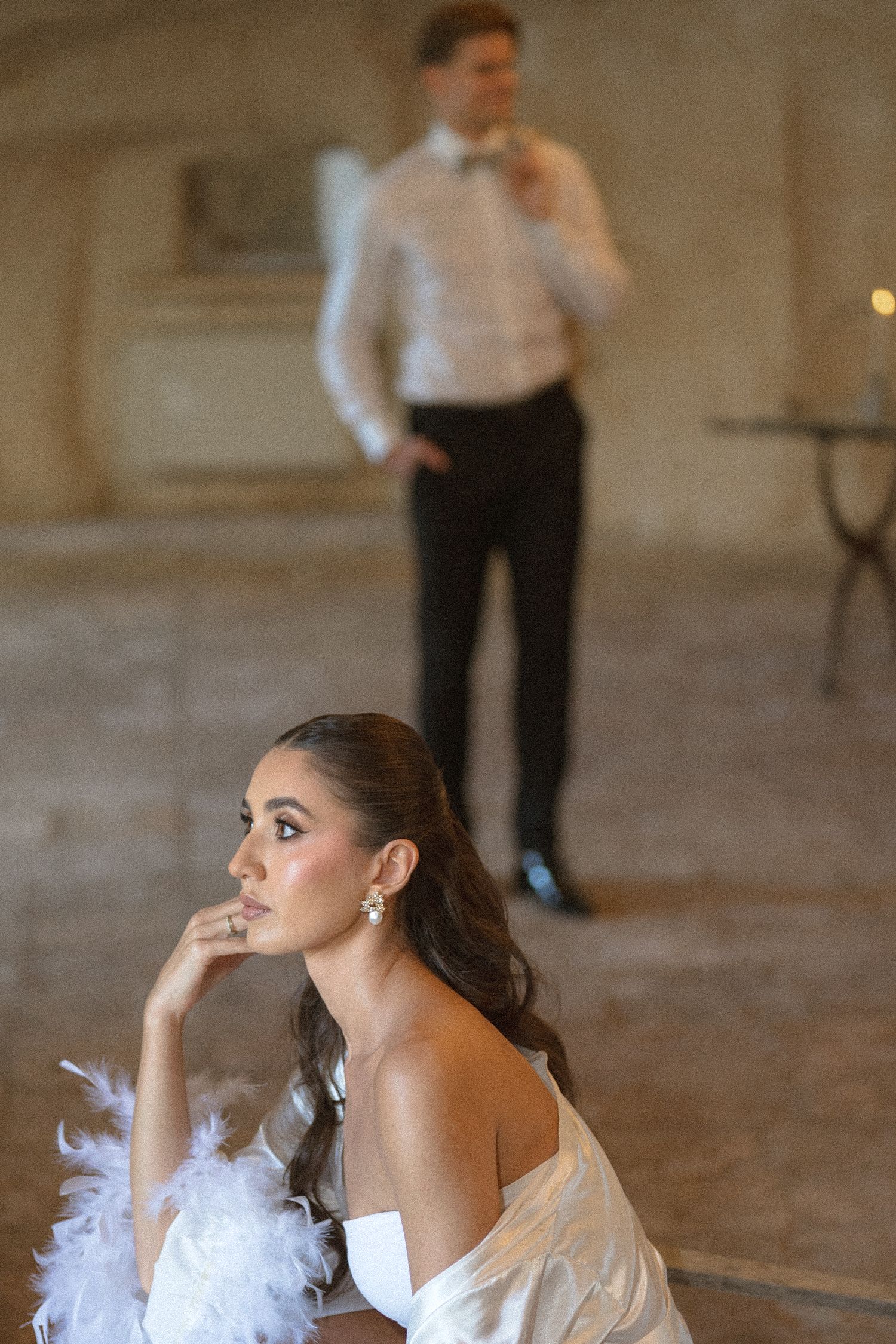 A woman in a white off-shoulder outfit with feather trim rests her chin on her hand while a groom in a bow tie stands blurred behind.