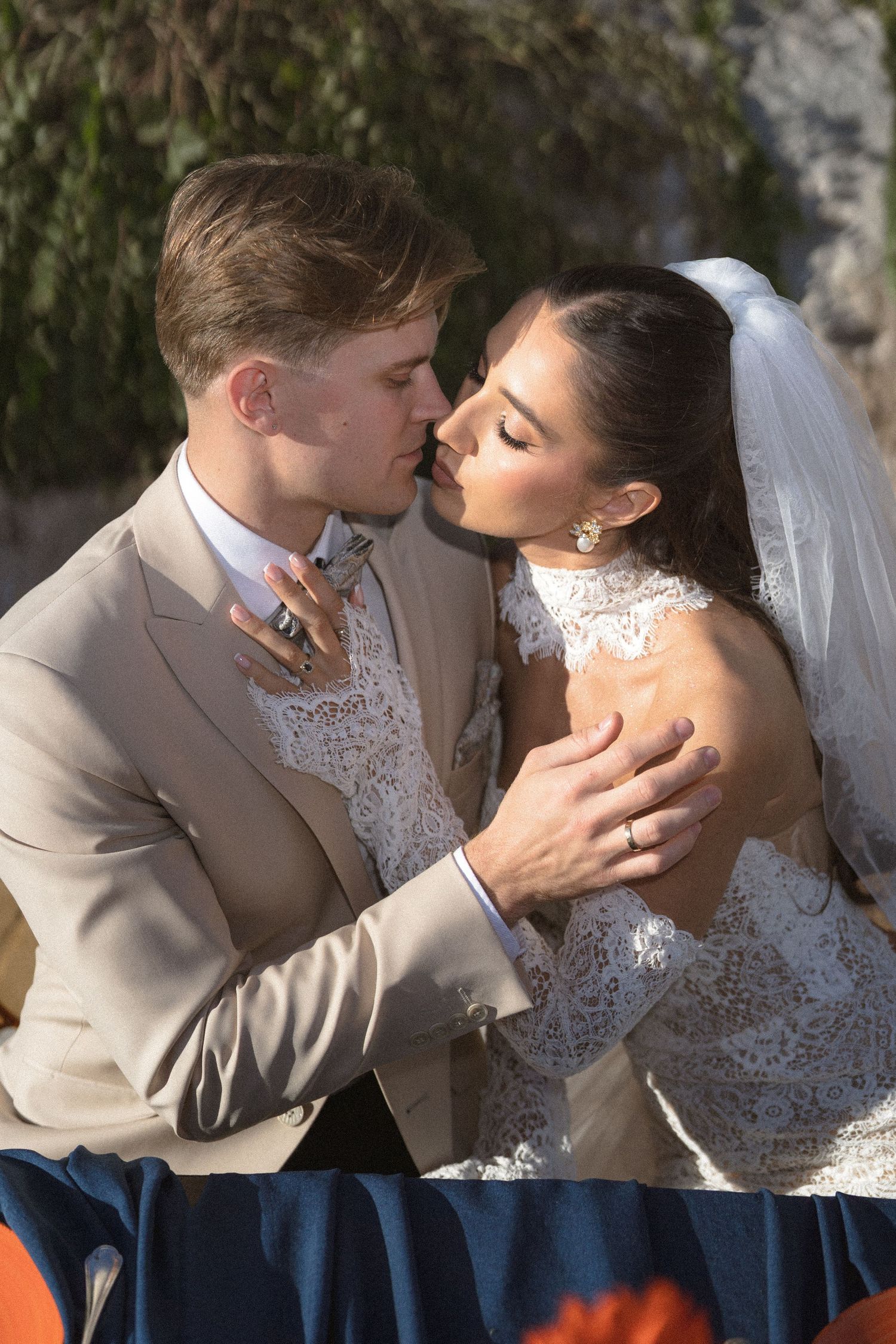A groom in a tan suit and a bride in a lace wedding dress and veil share an intimate moment, nearly kissing outdoors.