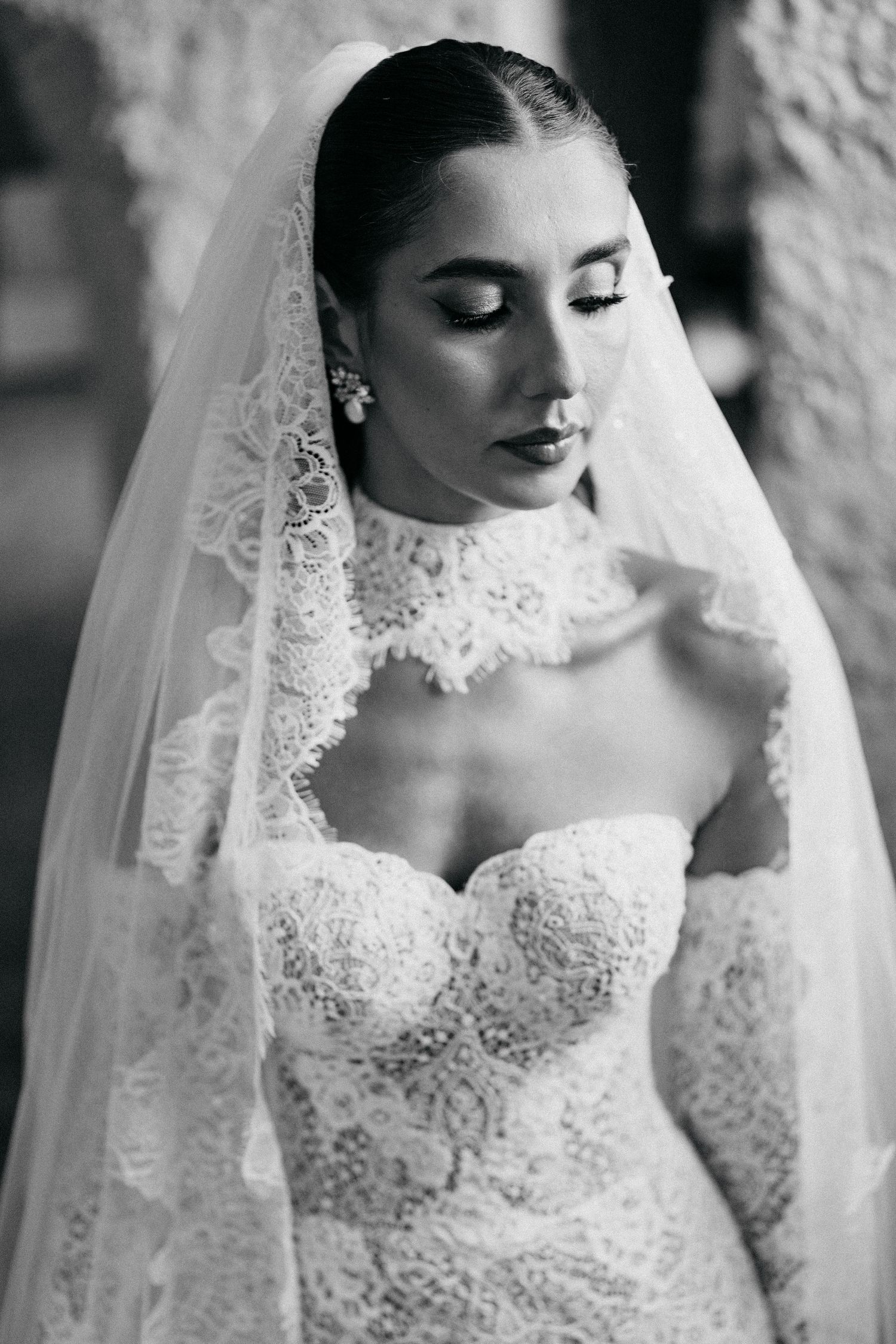 Black and white portrait of a bride with downcast eyes wearing a lace high-neck wedding gown and a lace-trimmed veil.