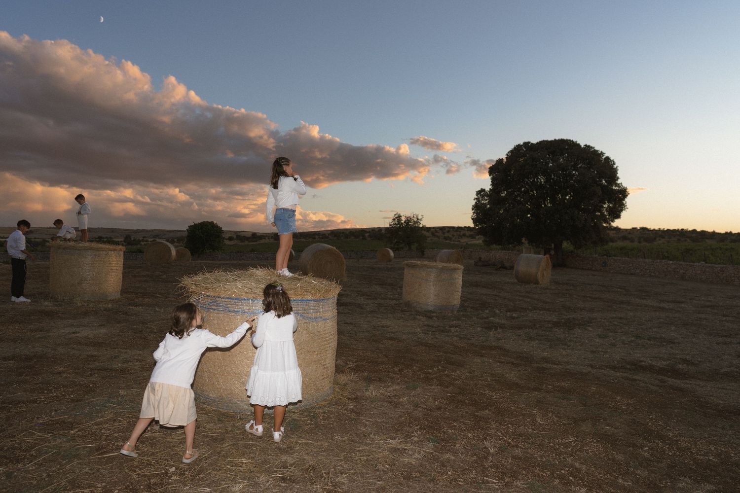 Children playing on round hay bales in a golden field at sunset, with a tree and dramatic clouds in the background.