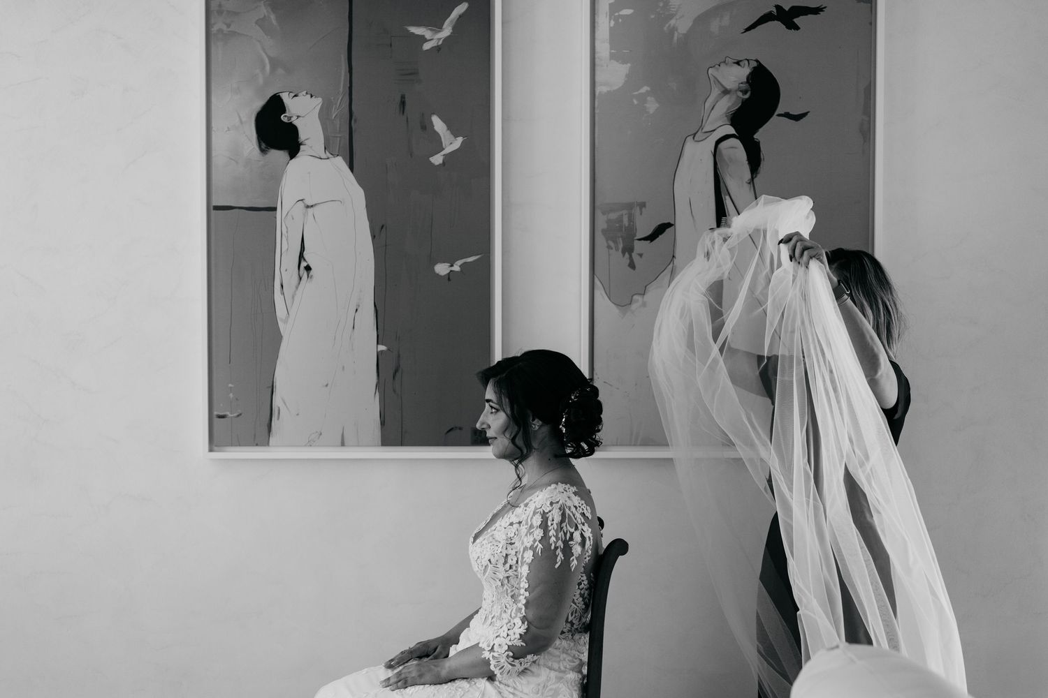 Black and white photo of bride in lace gown seated while bridesmaid arranges flowing veil before wedding paintings.