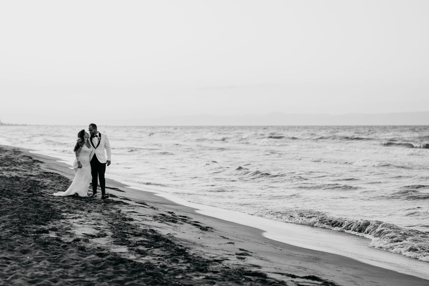 Black and white photo of newlyweds walking along ocean shoreline.