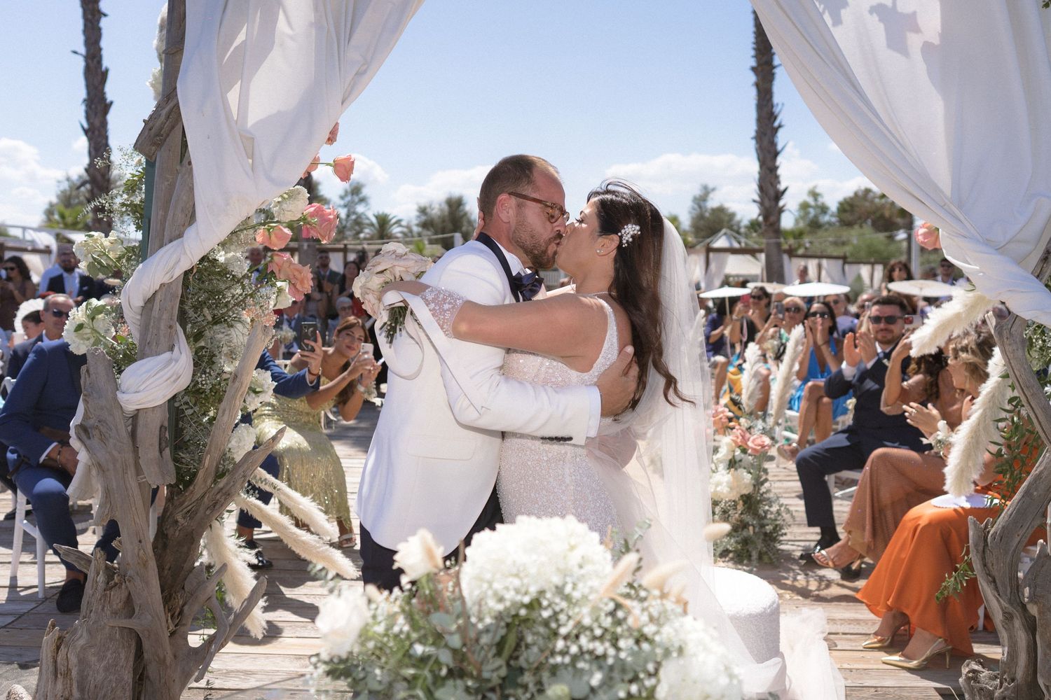 Beach wedding ceremony with flowing white fabric drapes and floral arrangements.