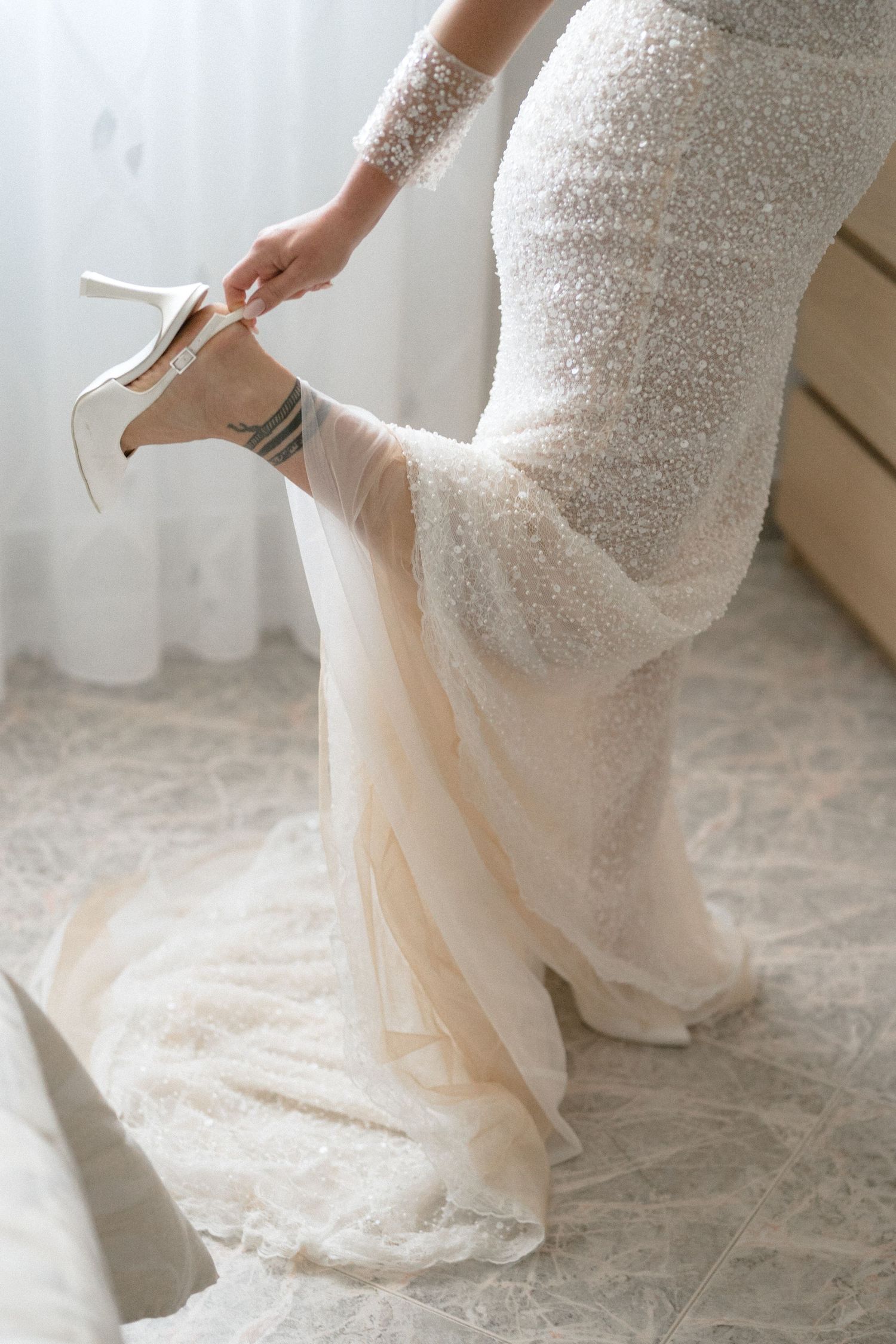 Close-up details of a beaded white wedding dress with flowing fabric on a light carpet.