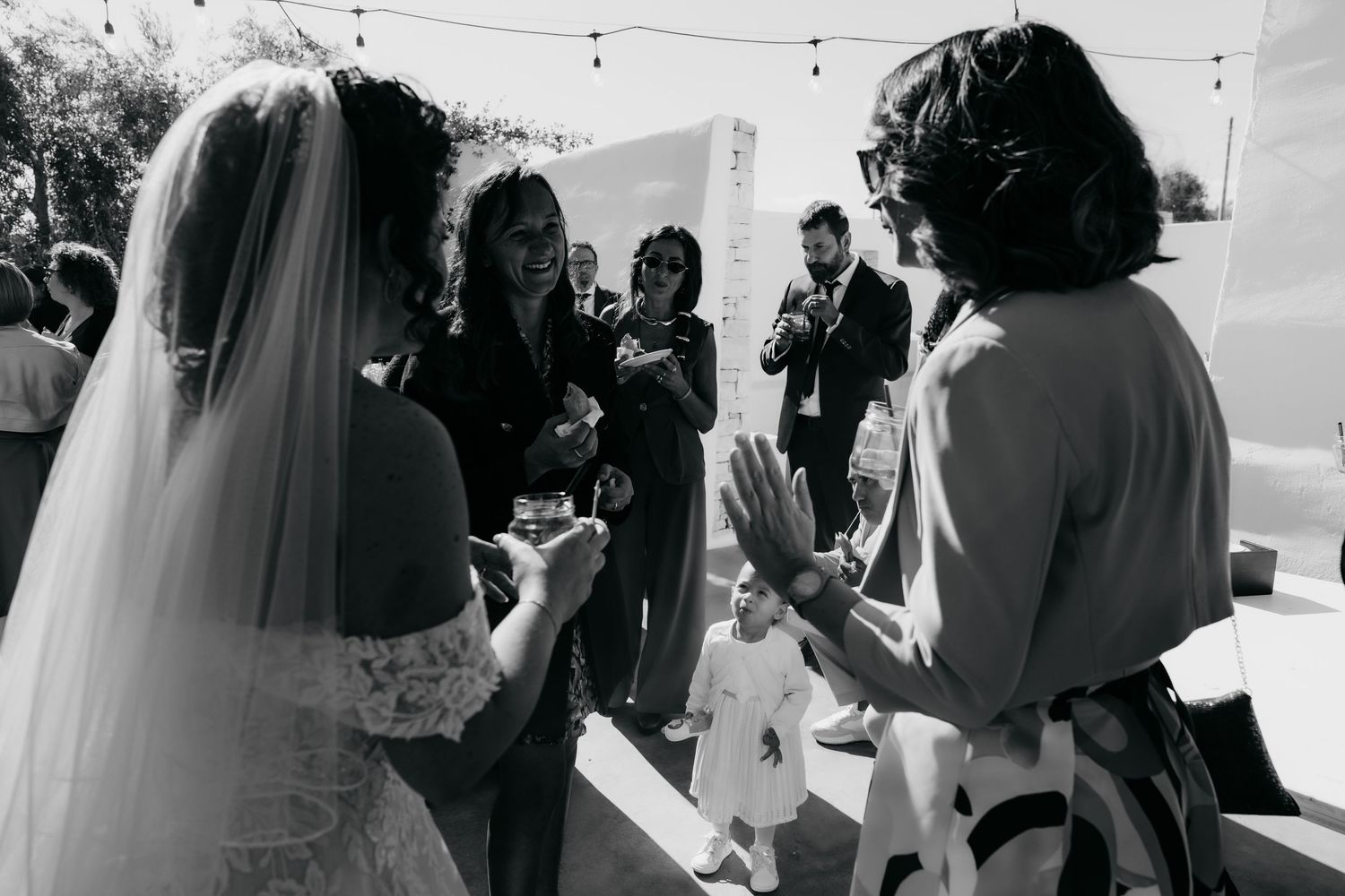 A bride in a white veil laughs with guests during an outdoor wedding reception in black and white photography.
