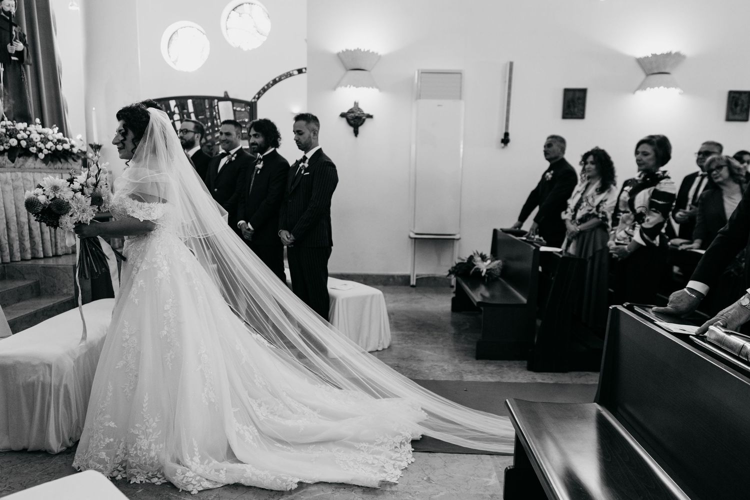 Bride in lace gown with long veil walks down church aisle with groom in black and white photo.