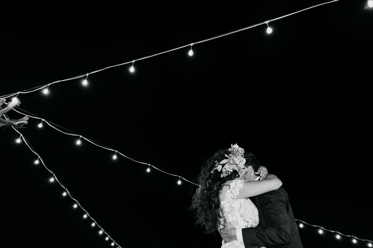 A couple embraces on the dance floor under romantic string lights at an outdoor evening celebration.