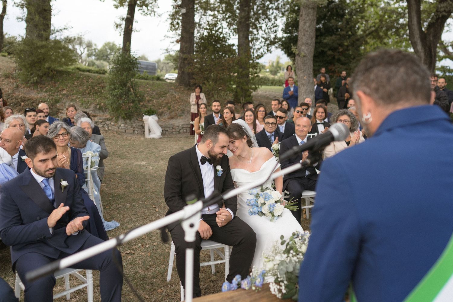 Outdoor wedding ceremony in a wooded setting with guests seated on white chairs watching the couple exchange vows.