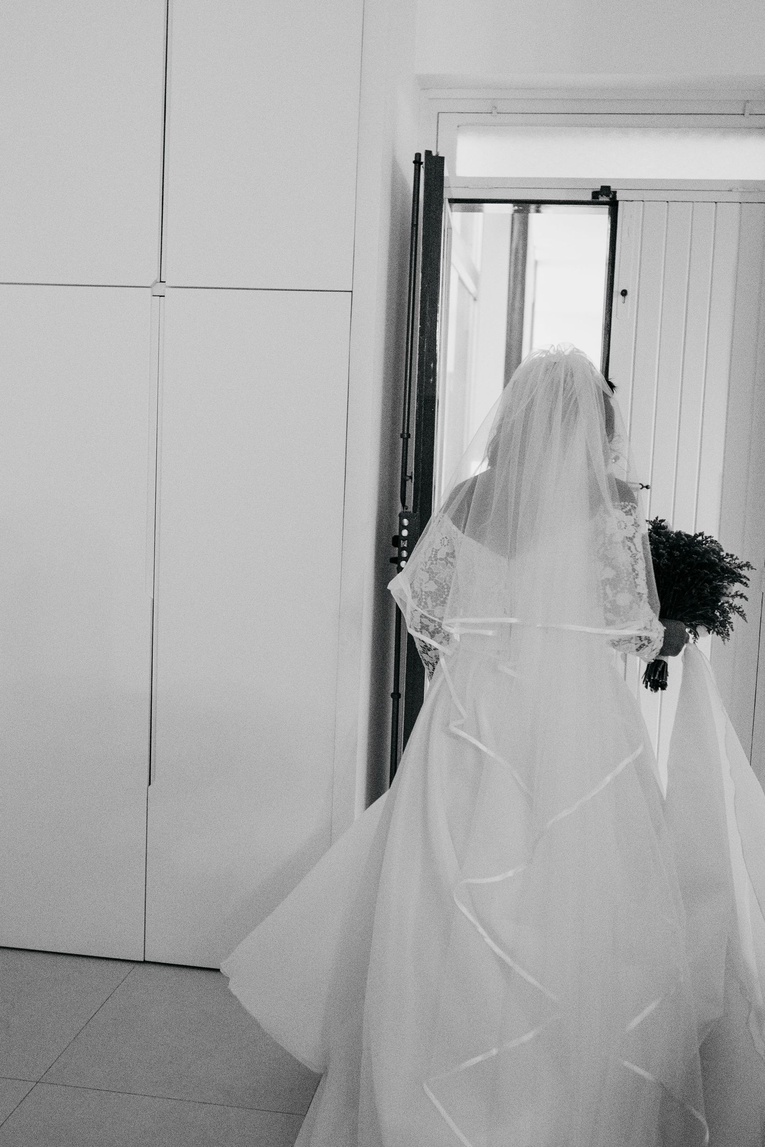 A black and white photo of a bride in full wedding attire viewed from behind.