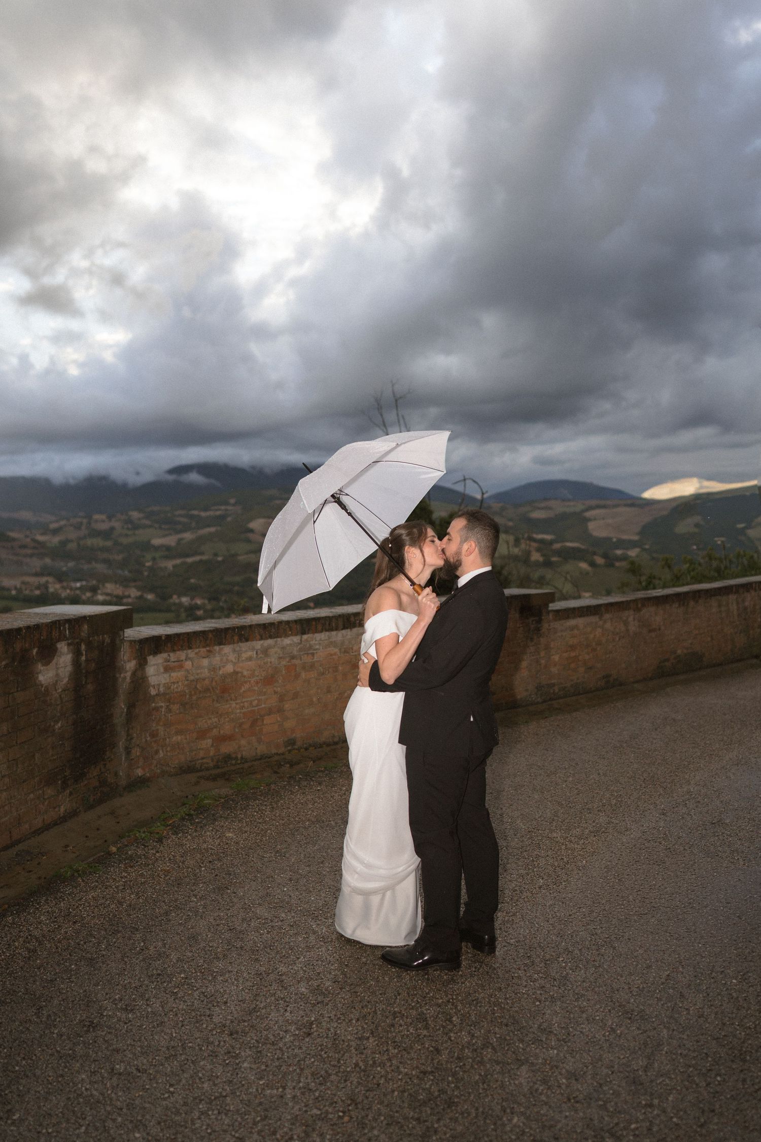 Couple sharing romantic moment under white umbrella against dramatic cloudy mountain backdrop.