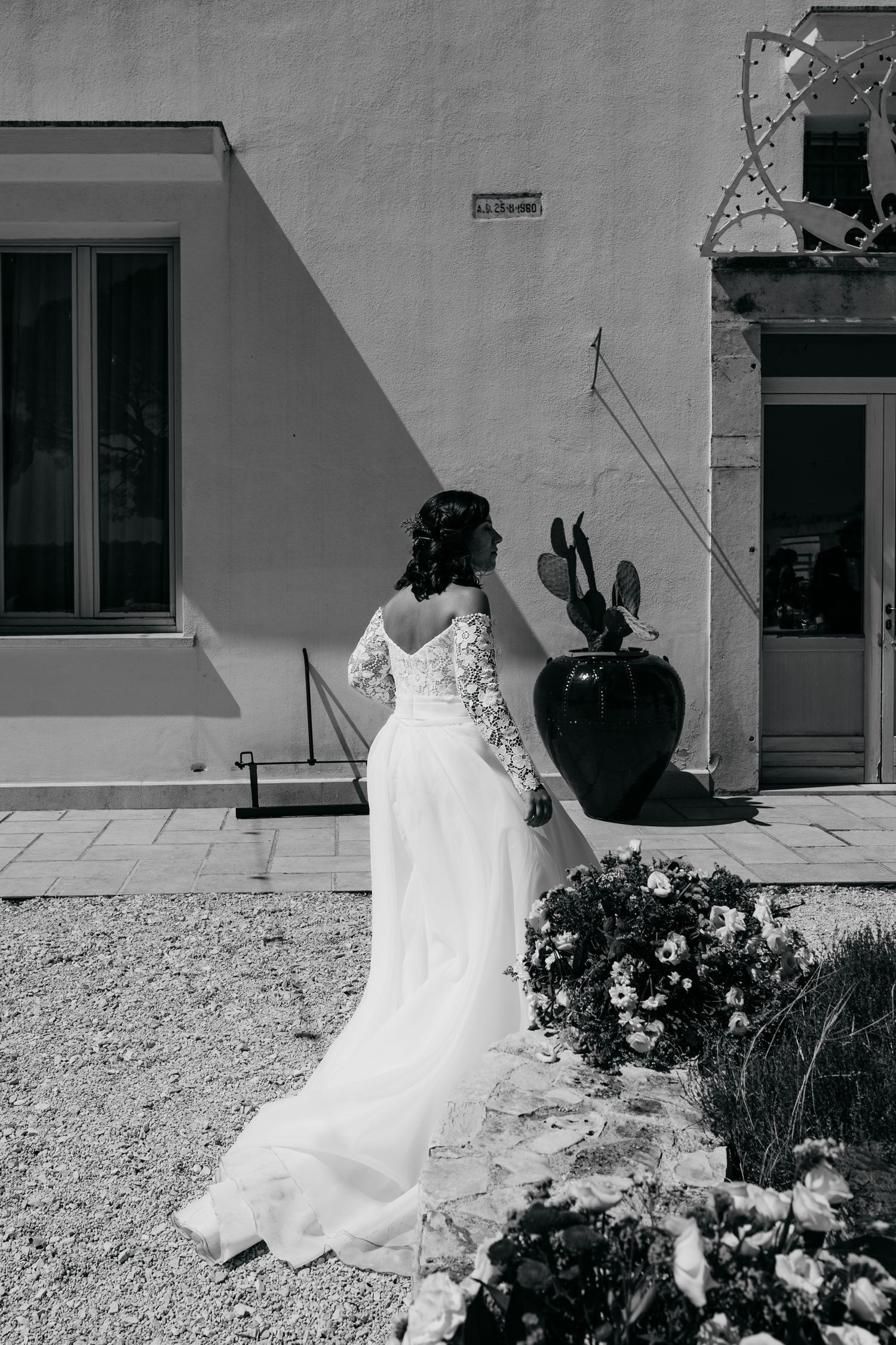 Black and white photo of a wedding dress flowing in the breeze against a textured wall and garden.