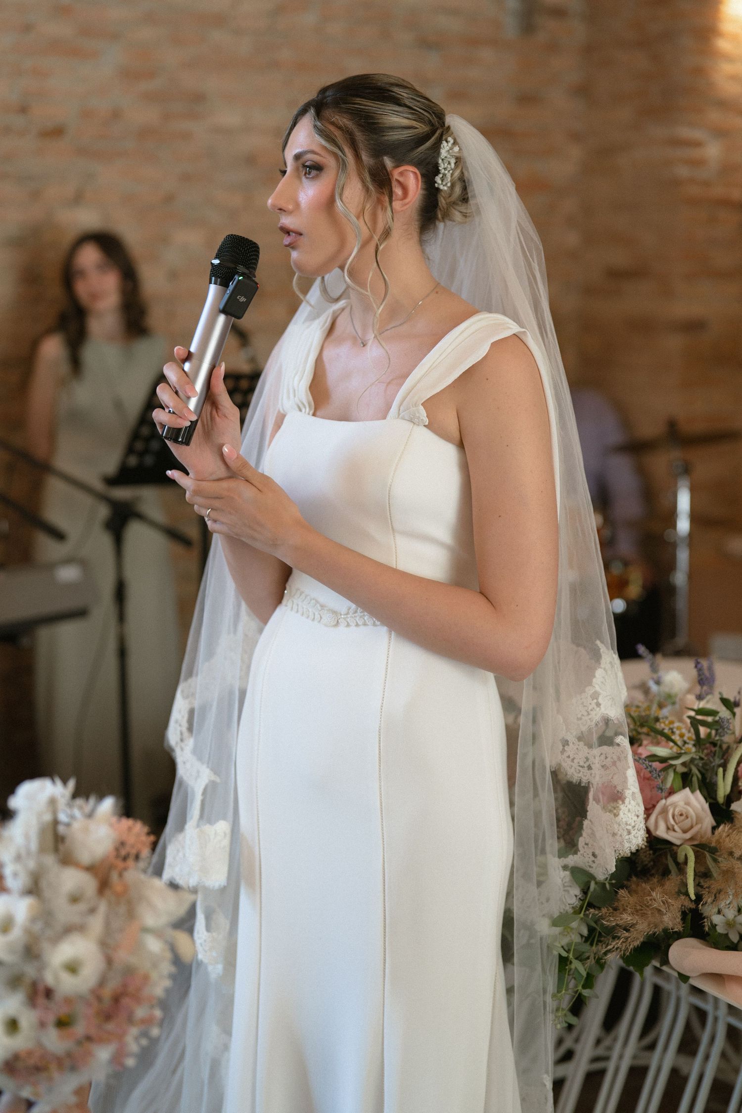 Bride in white wedding dress giving an emotional speech at a rustic brick-walled reception venue.