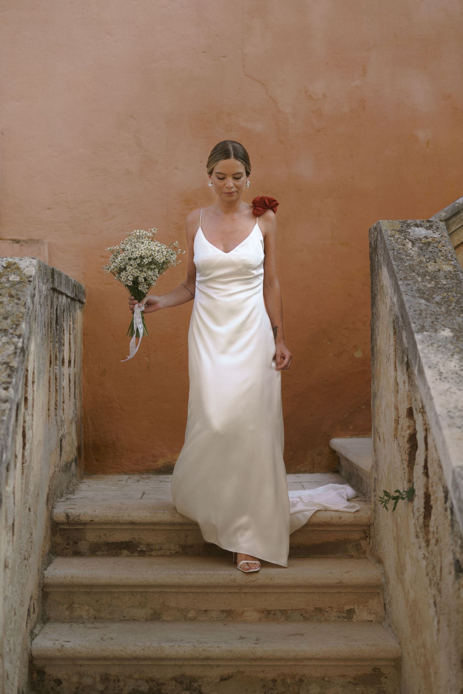 A bride in a simple white dress descends stone steps while holding a delicate bouquet of flowers.