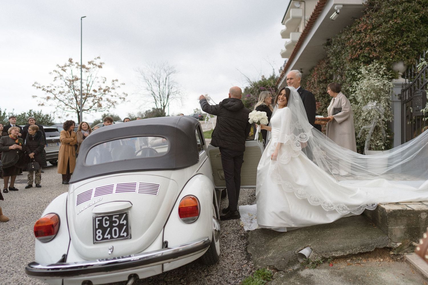 Bride in white gown arriving at wedding venue in a classic white Volkswagen Beetle car.