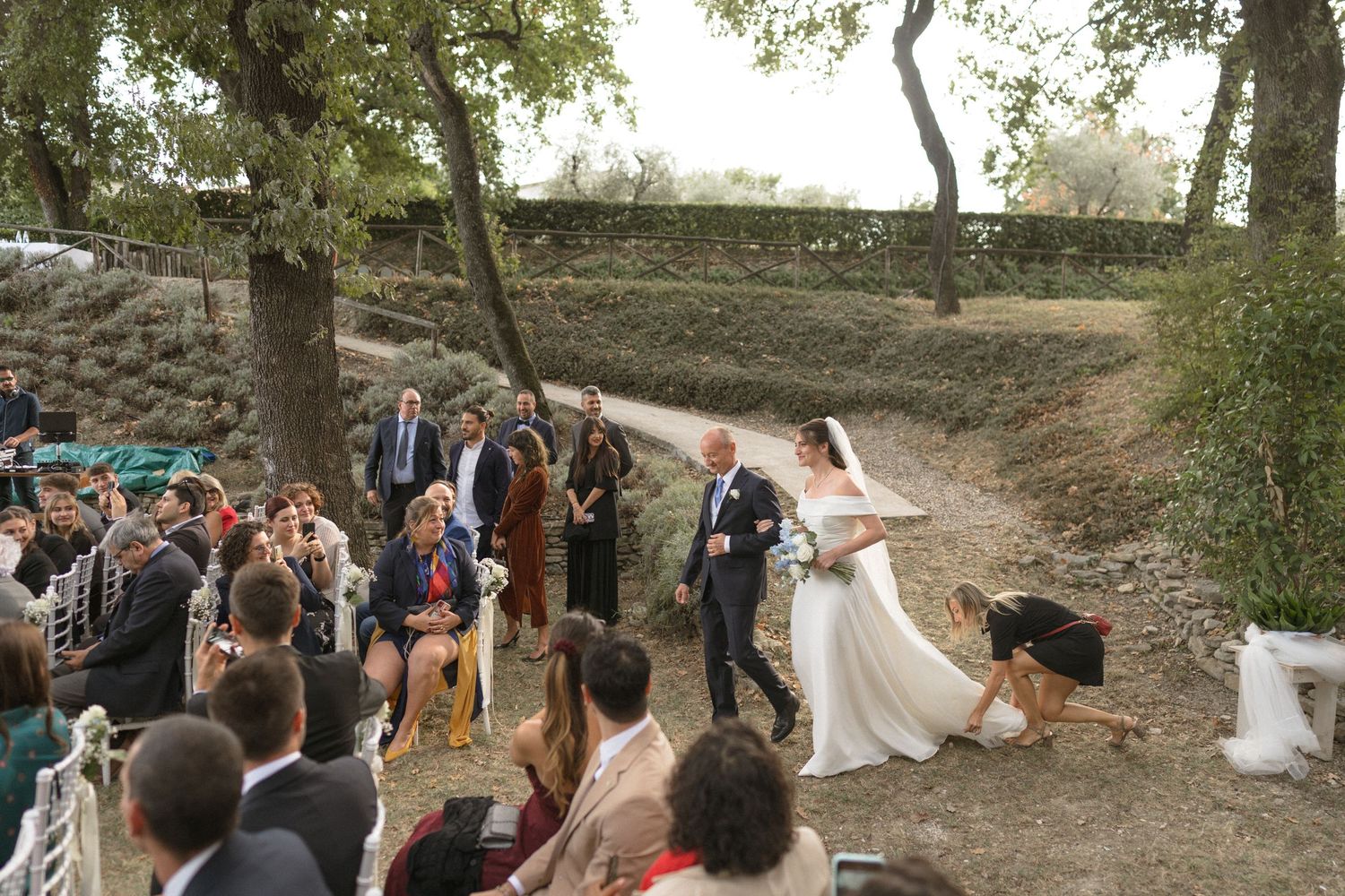 Panoramic wedding celebration scene with guests gathered under trees in a rustic outdoor setting.