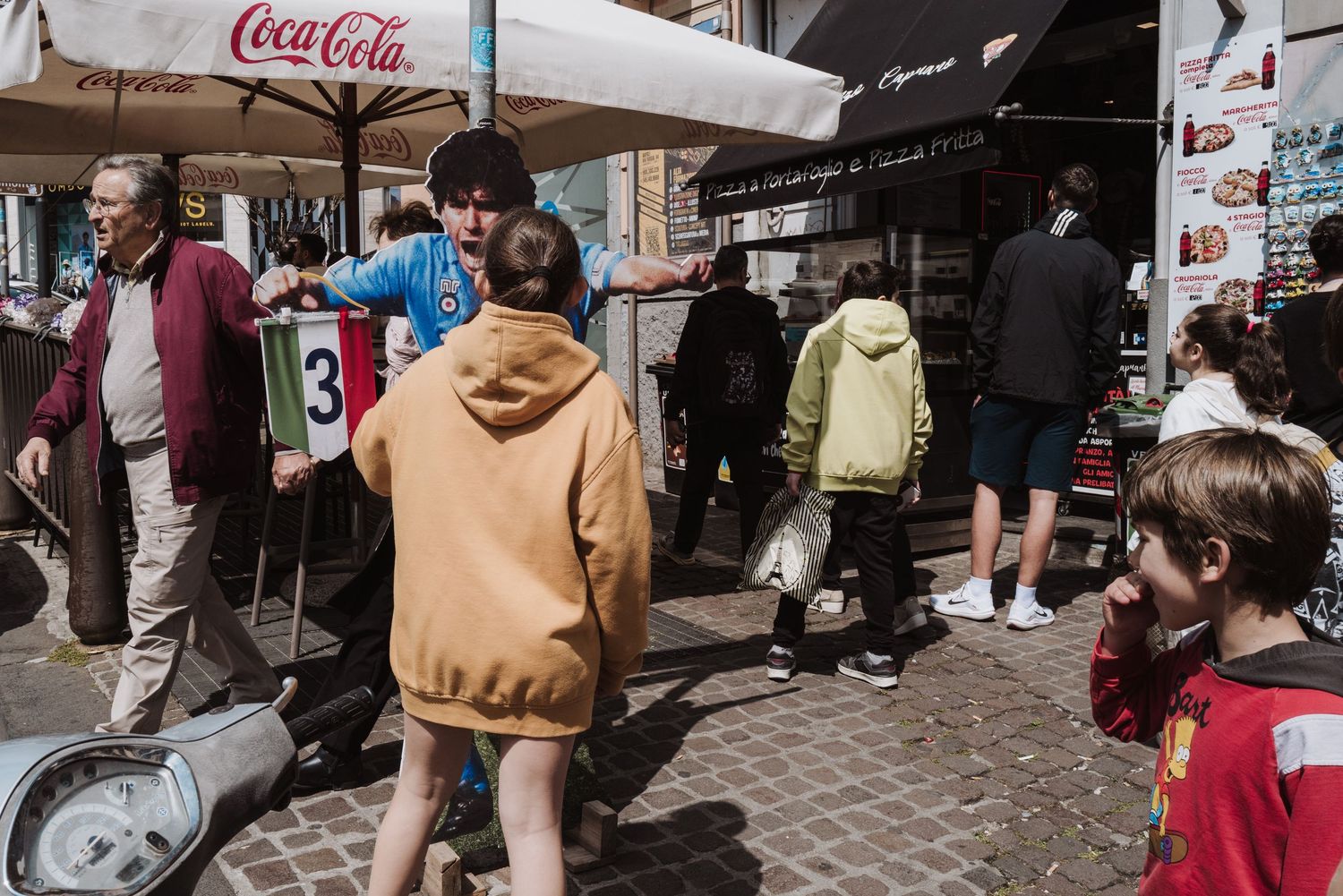 People gather under a Coca-Cola umbrella on a busy cobblestone street during daytime.