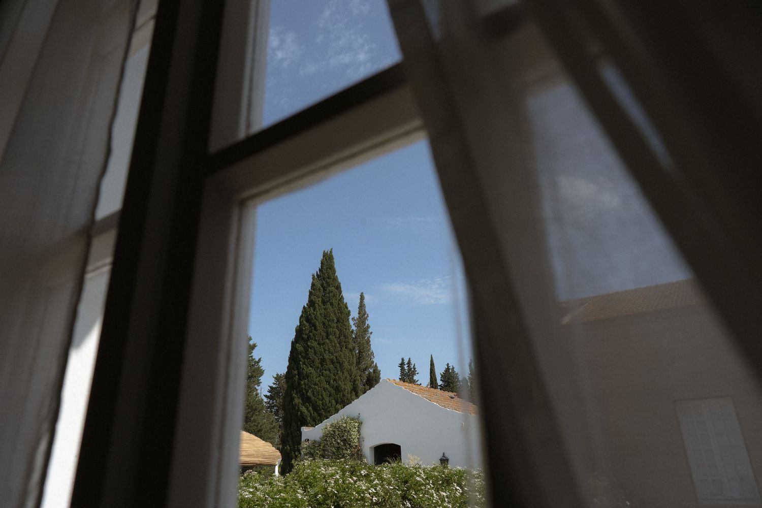 A wooden window frame with white curtains looks out onto cypress trees and a Mediterranean style building exterior.