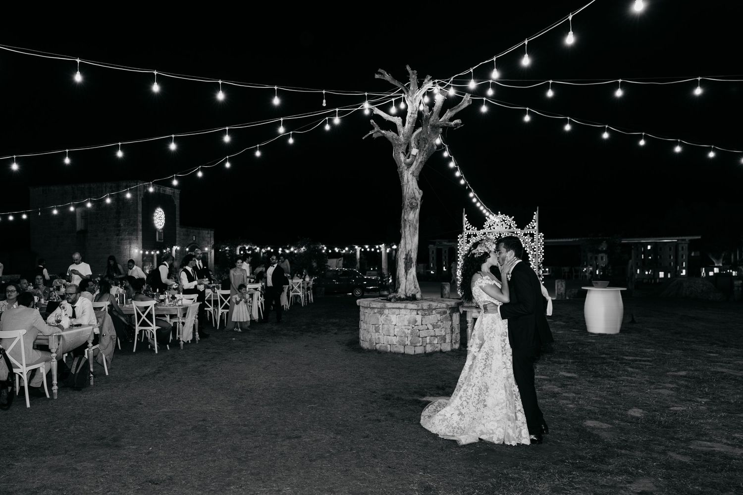 Outdoor evening wedding reception under twinkling string lights with white chairs and tables arranged on a lawn.