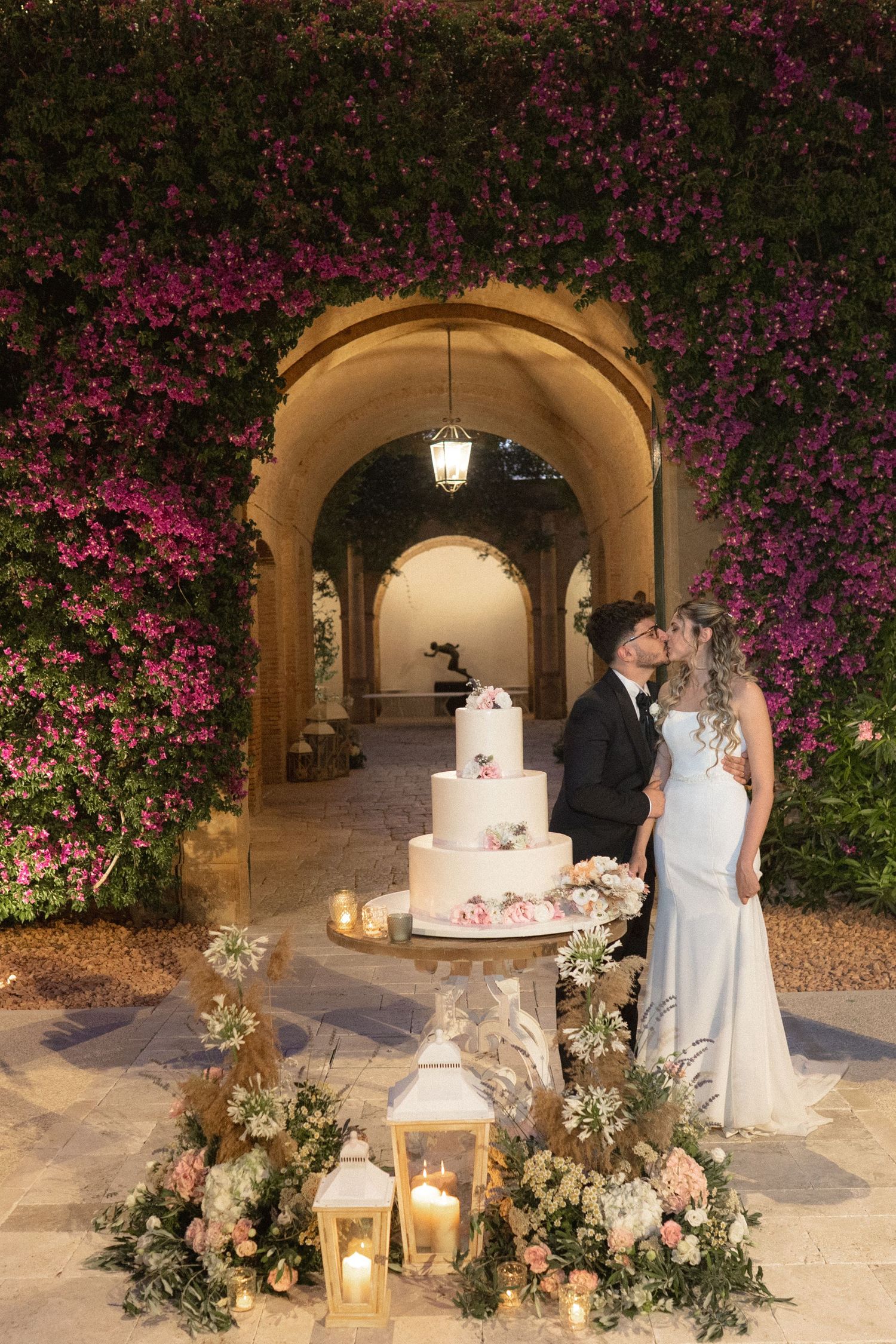 Elegant wedding cake display with lanterns and floral arrangements under an archway at night.