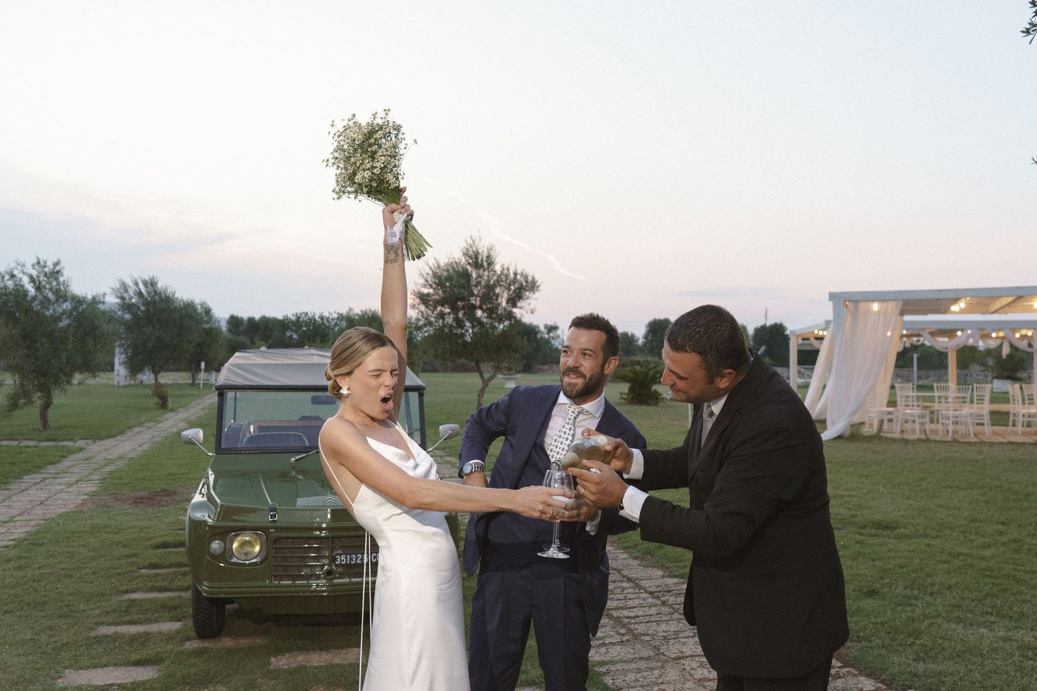Wedding celebration outdoors with vintage Land Rover in background at sunset.
