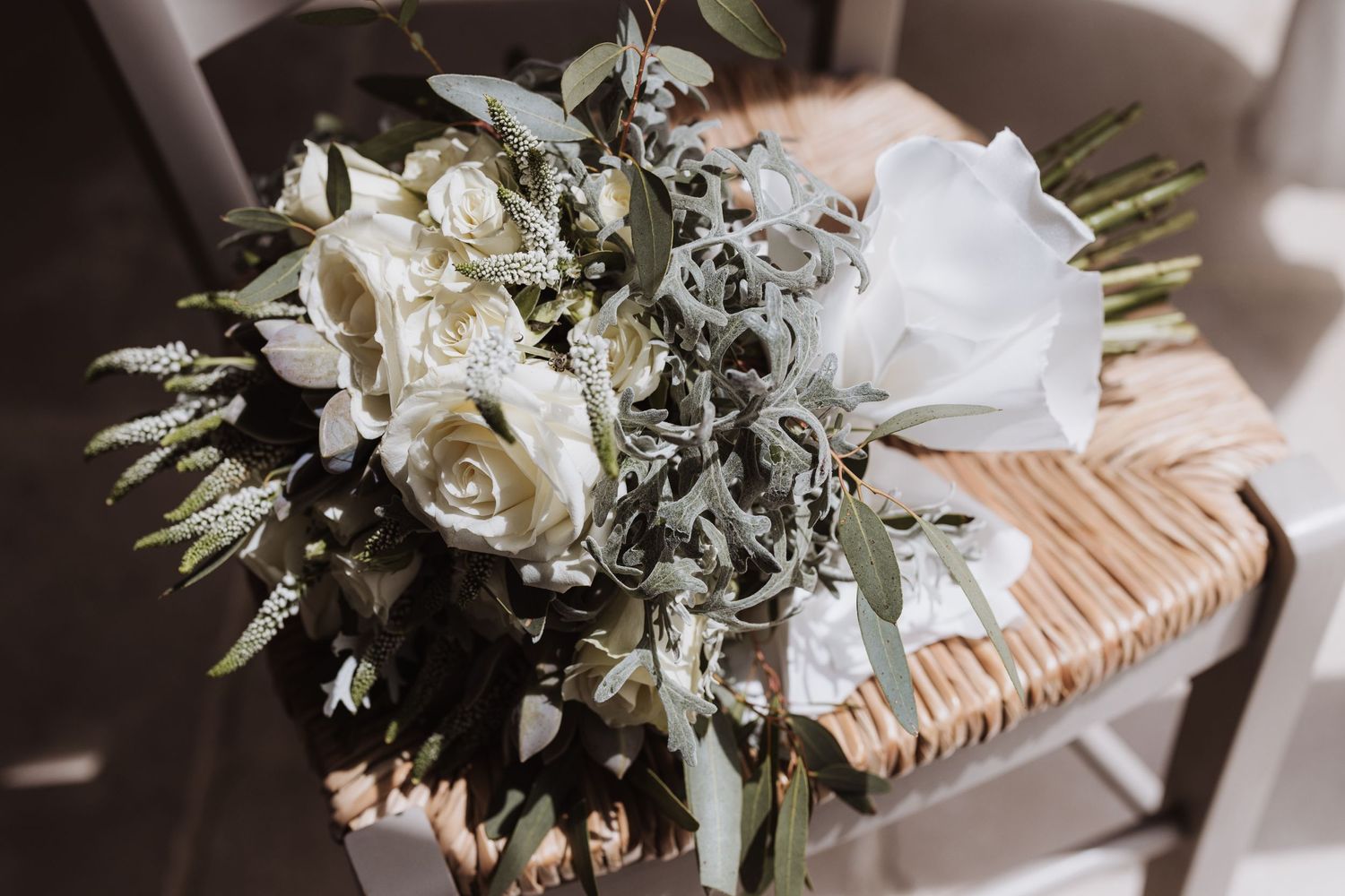 A classic white wedding bouquet with roses and greenery rests on a woven chair.