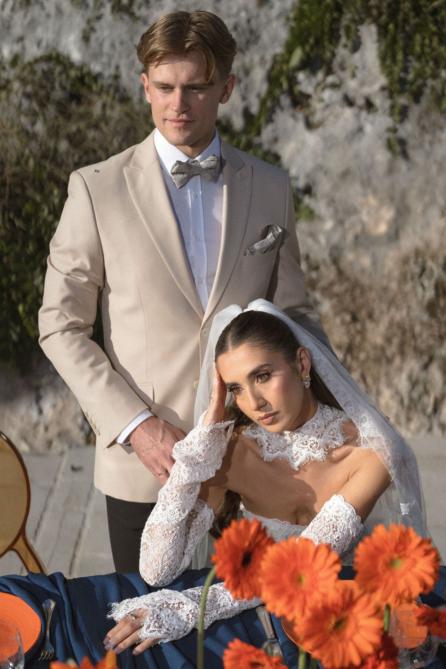 Groom in a beige blazer and bow tie stands behind a bride in a lace gown and veil seated at a table with orange gerbera daisies.