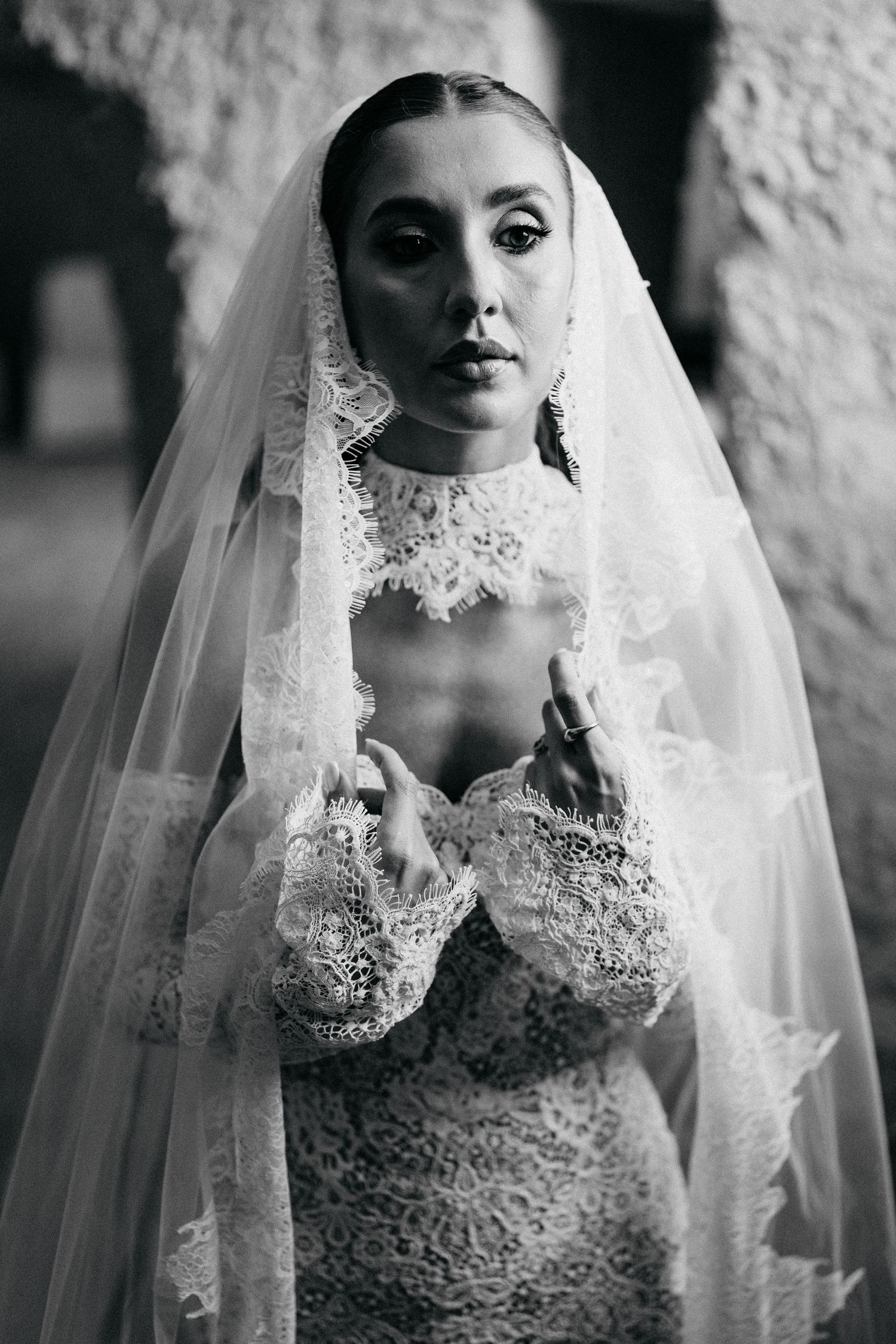 Black and white portrait of a bride in a high-neck lace wedding dress and sheer veil touching her collar near a stone wall.