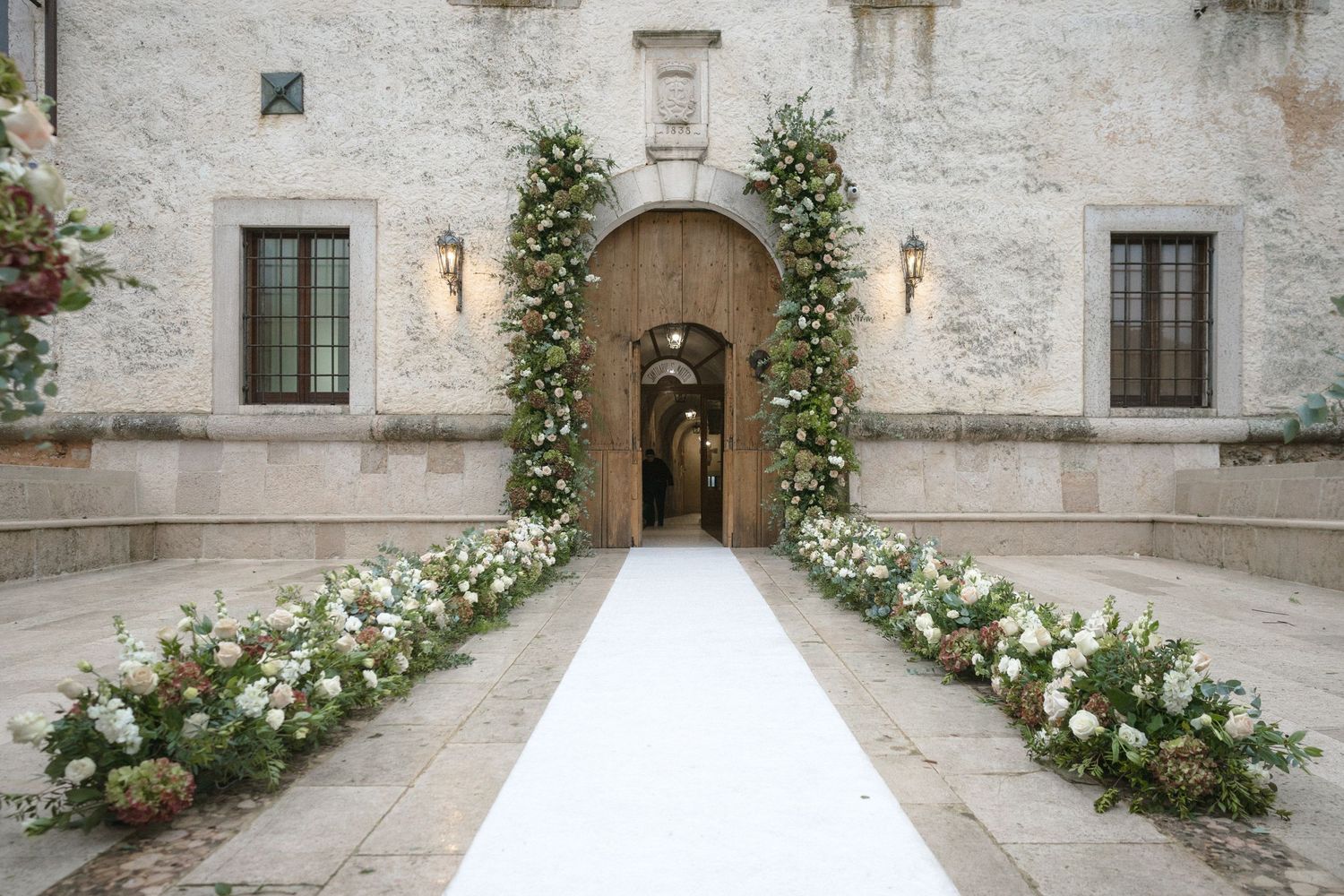 Elegant wedding venue entrance with white floral arrangements lining a white aisle runner.