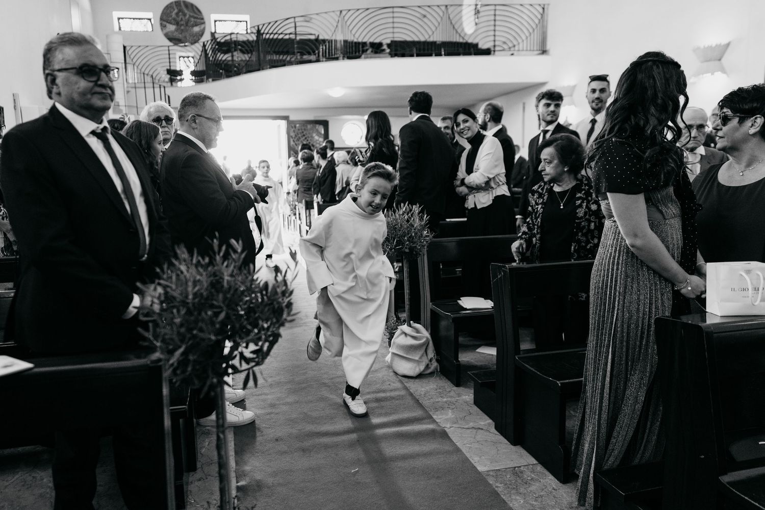 Child in white runs down church aisle during wedding ceremony in black and white photo.