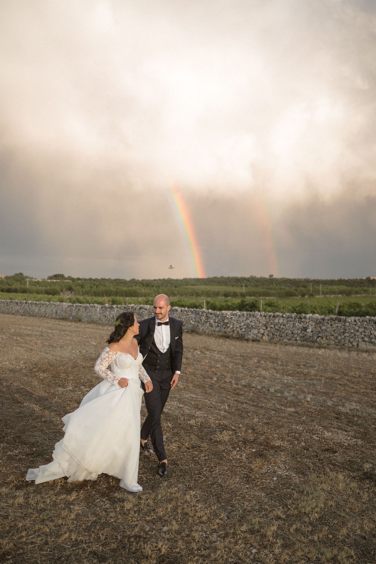 A couple embraces on a rustic dirt path with a stone wall and olive trees in the background at sunset.