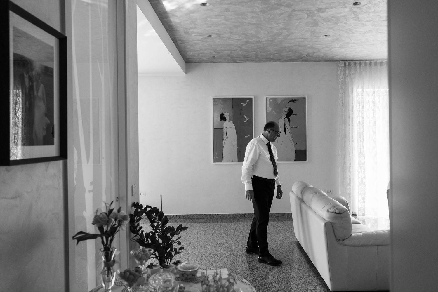 Black and white photo of a man in a suit standing in an elegant room with artwork on the walls and flowers in the foreground.