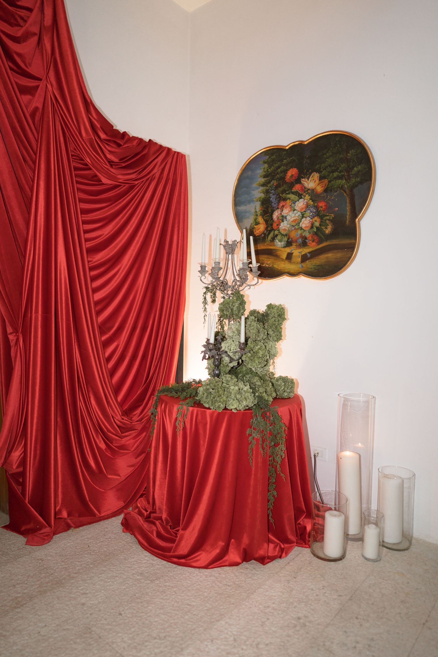 Elegant wedding decor with red satin curtain, candelabra, greenery centerpiece, and white candles.
