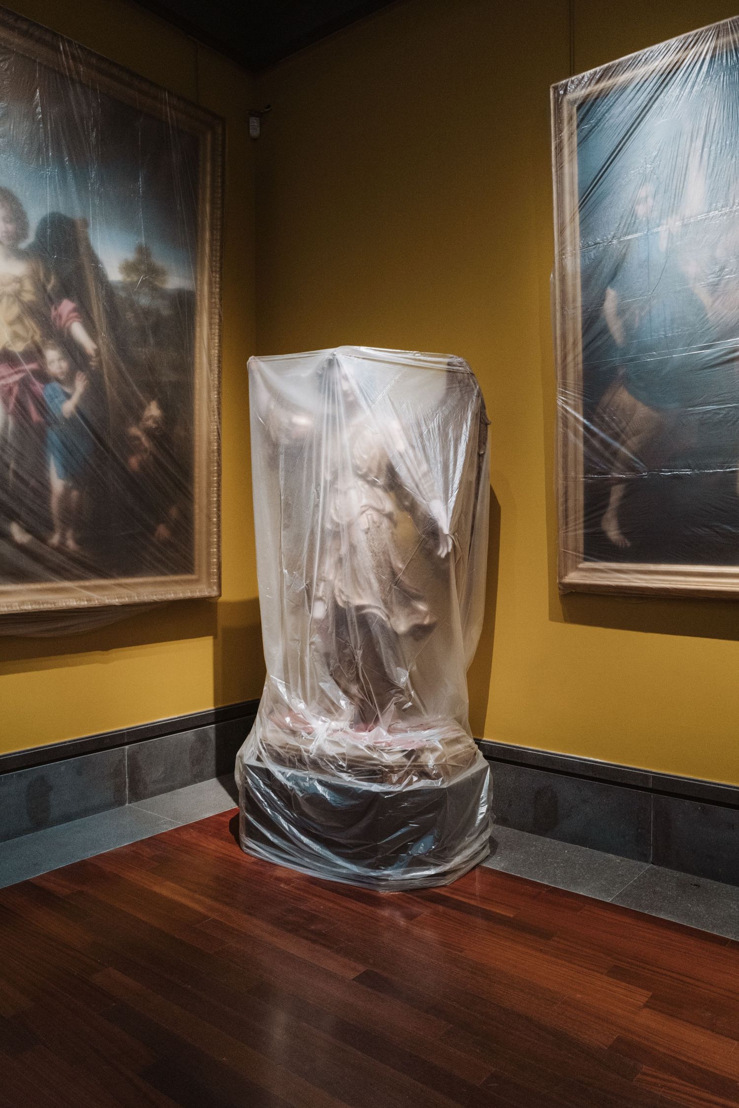 Statue covered in protective plastic wrap displayed between two paintings in museum gallery with yellow walls.