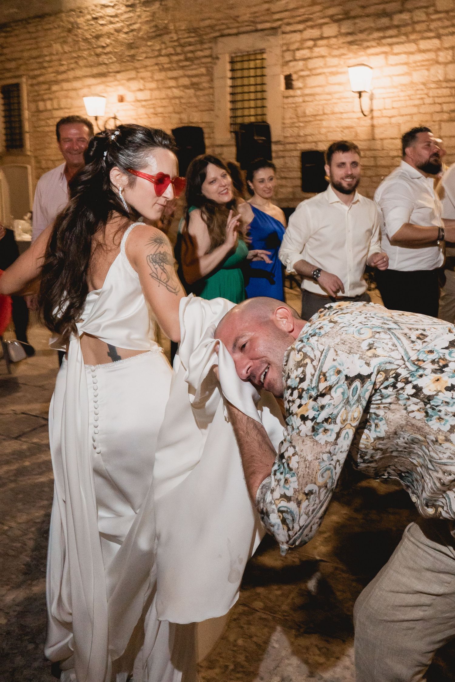 Wedding guests dance energetically at a reception with exposed brick walls and warm lighting.