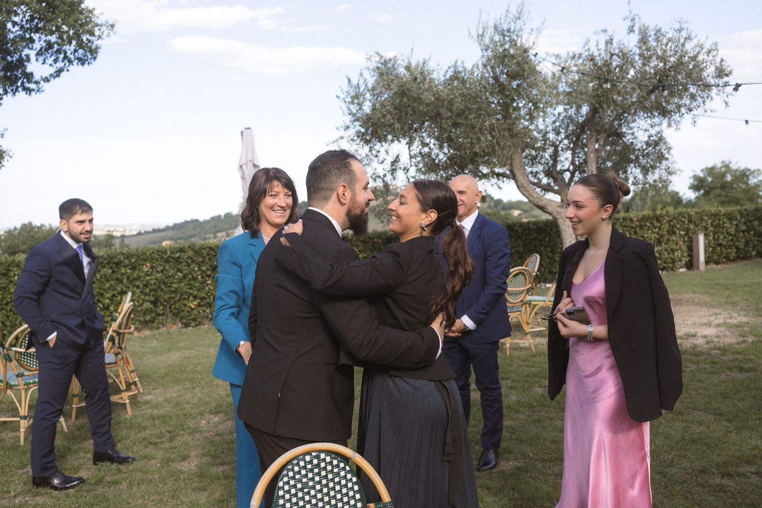 Wedding guests gathered outdoors among olive trees during a daytime celebration.
