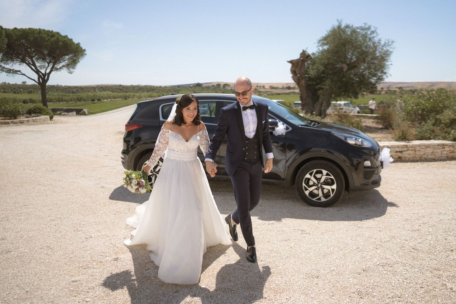 Newlyweds walking together on sunny path with car in background.