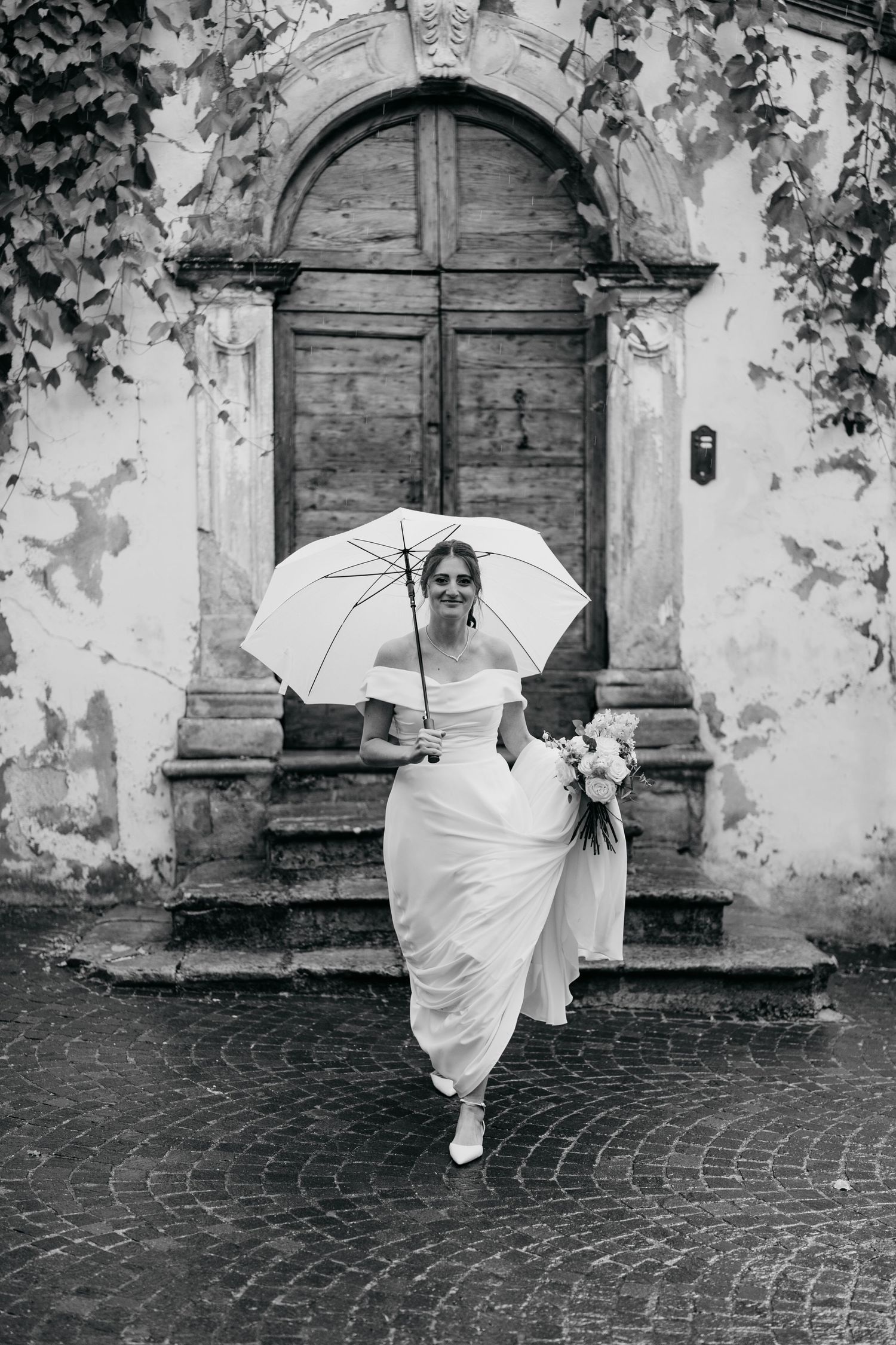 Black and white wedding photos showing a white umbrella against a rustic stone wall and cobblestone ground.