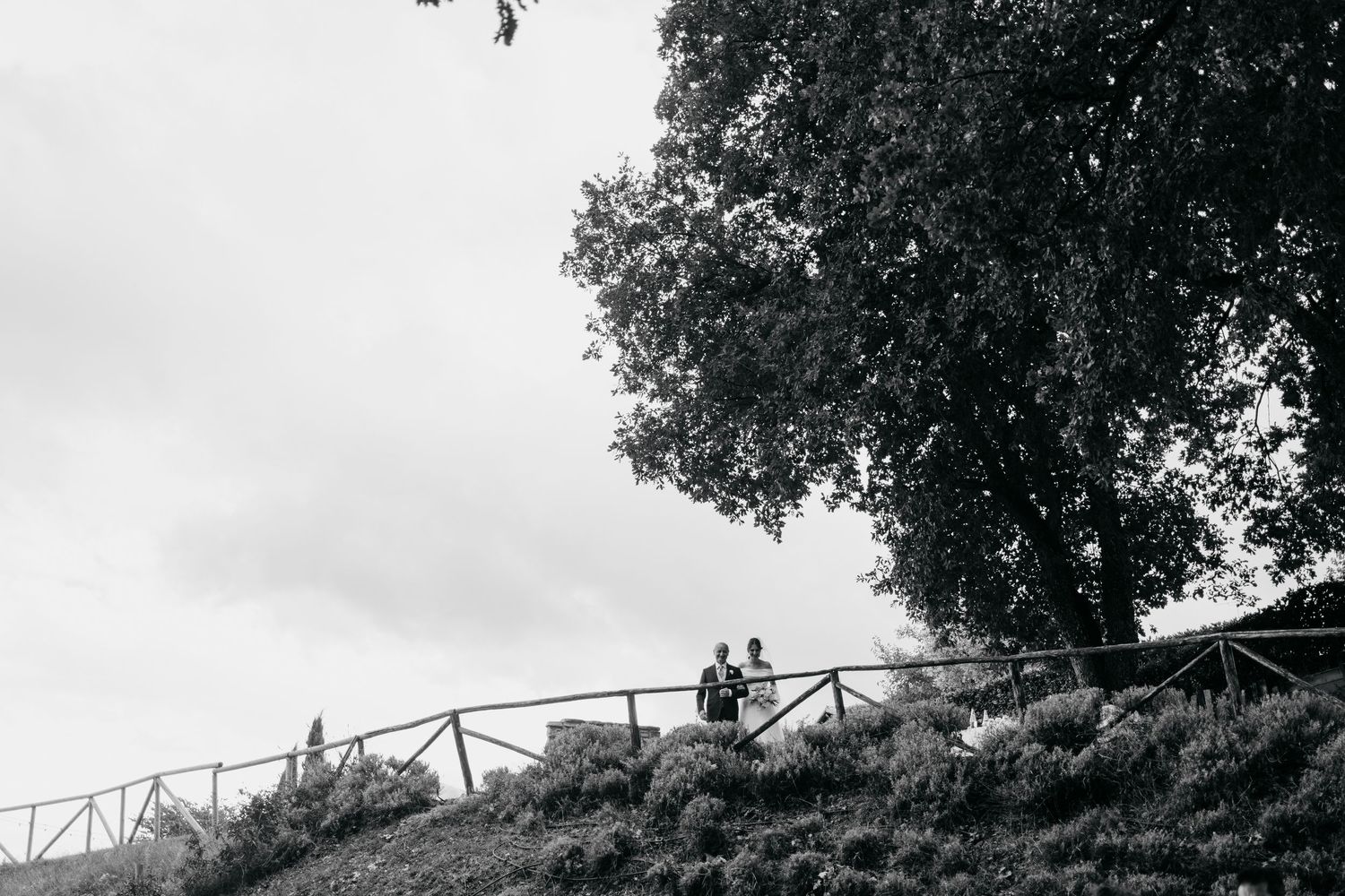 A couple stands on a rustic wooden bridge surrounded by lush greenery and trees in a romantic outdoor setting.